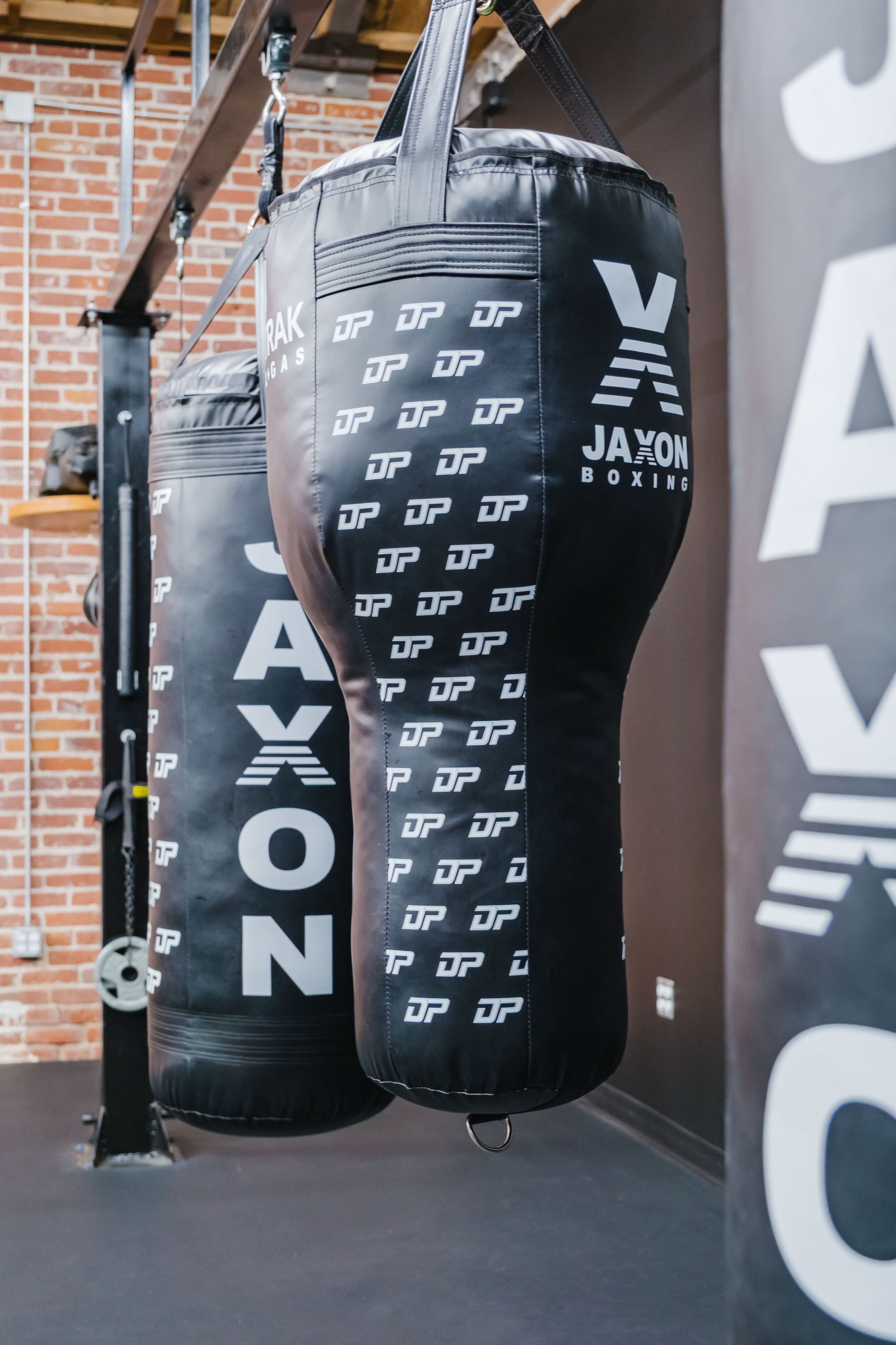 The image shows two black boxing bags hanging from a gym ceiling, with one prominently displaying the name "JAXON" and a logo.