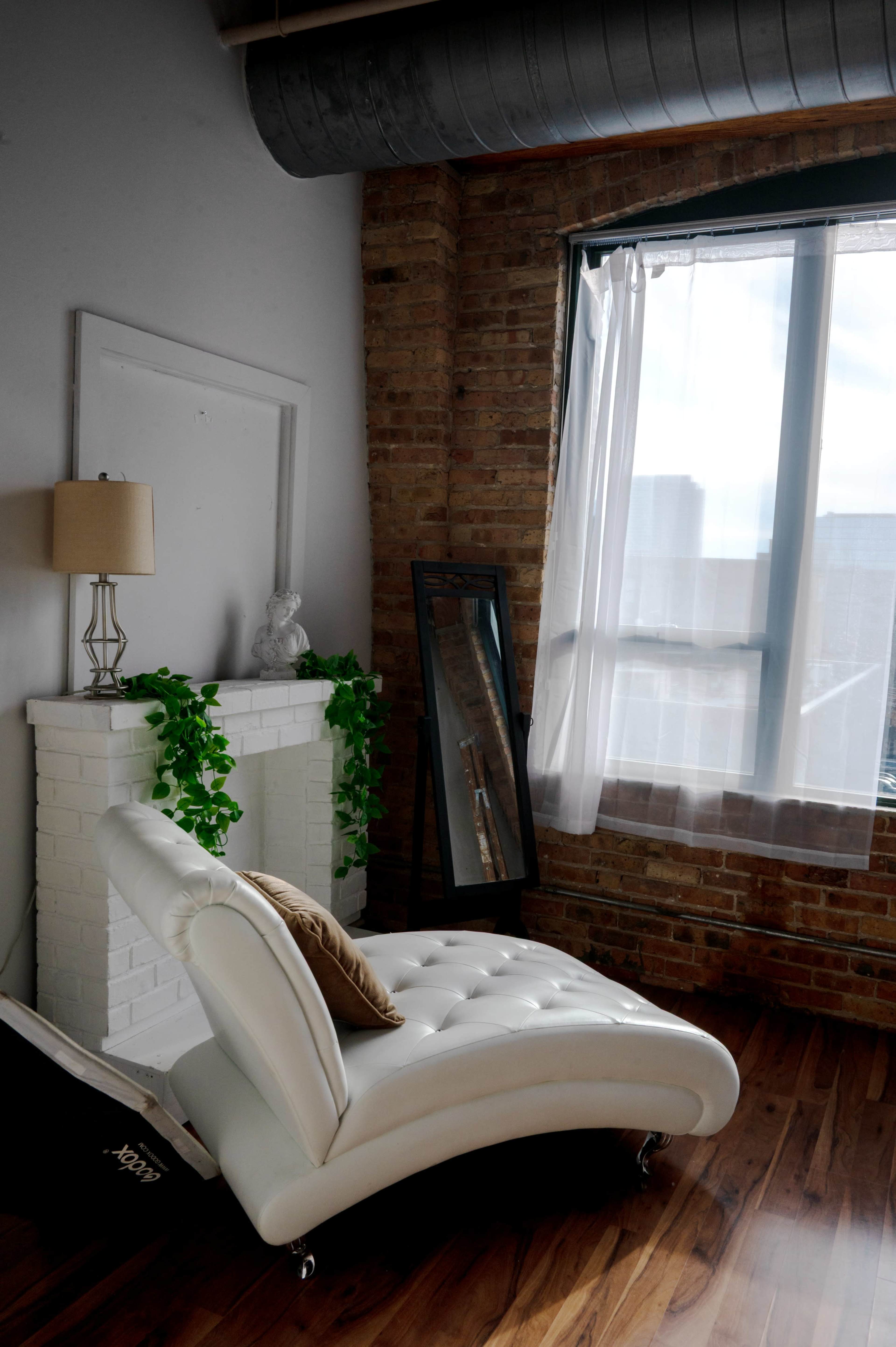 The image shows a modern living space featuring a white chaise lounge positioned next to a brick wall, a faux fireplace, a tall mirror, a lamp, and a window with sheer curtains.