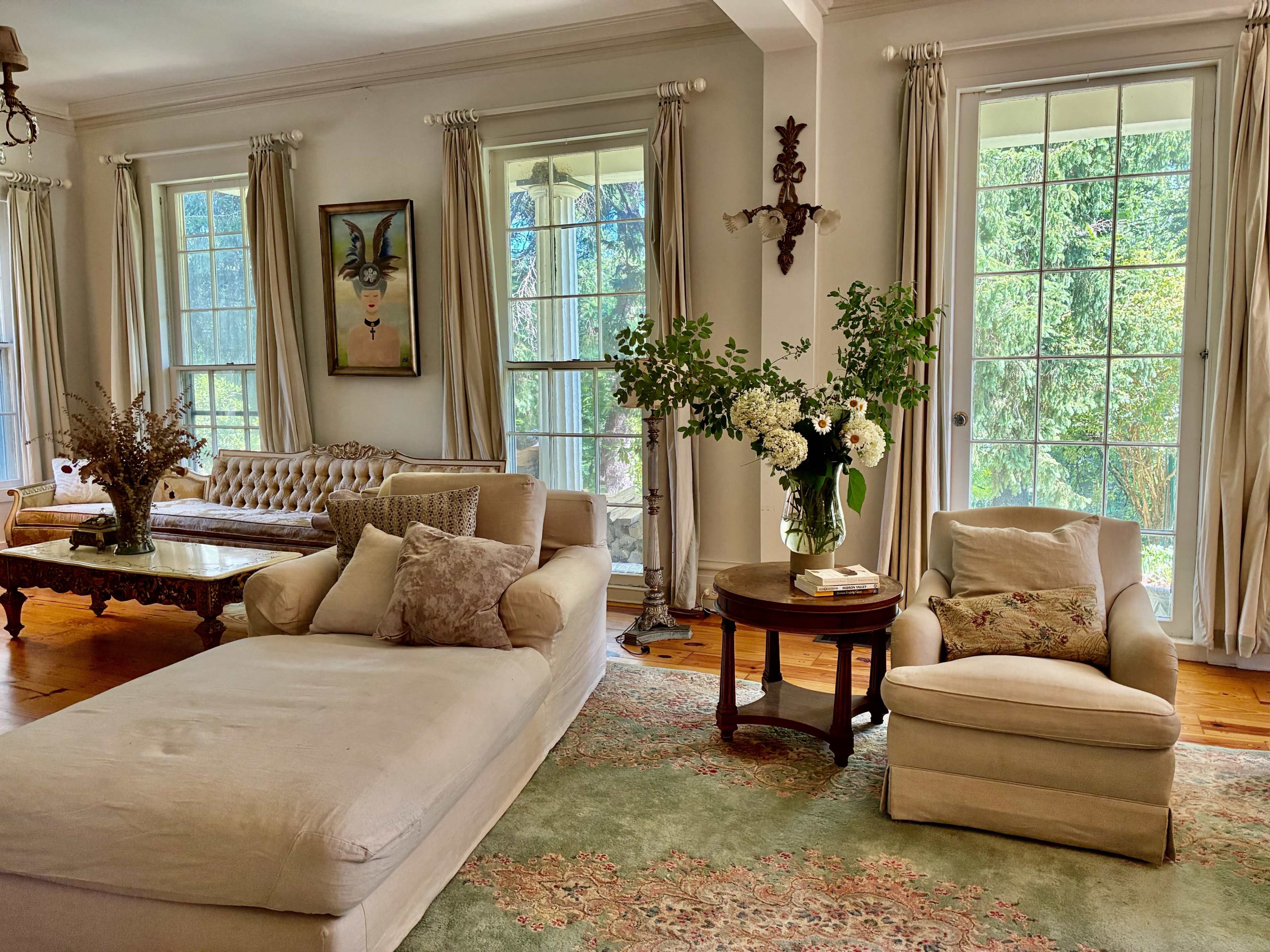 The living room features a large light-colored sectional sofa, a round table with a floral arrangement, and several large windows allowing natural light to illuminate the space.