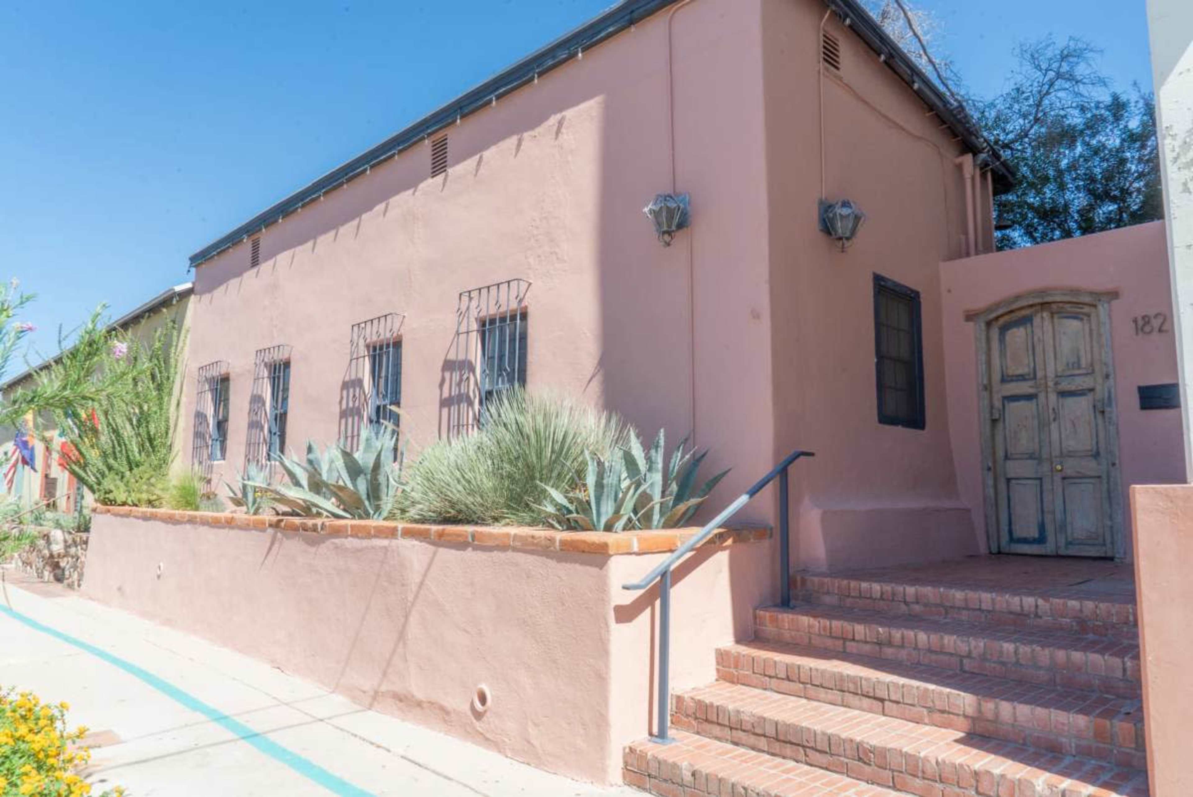 The image shows a single-story pink adobe building with a wooden door, blue trim, and several square windows, surrounded by low greenery and brick steps.