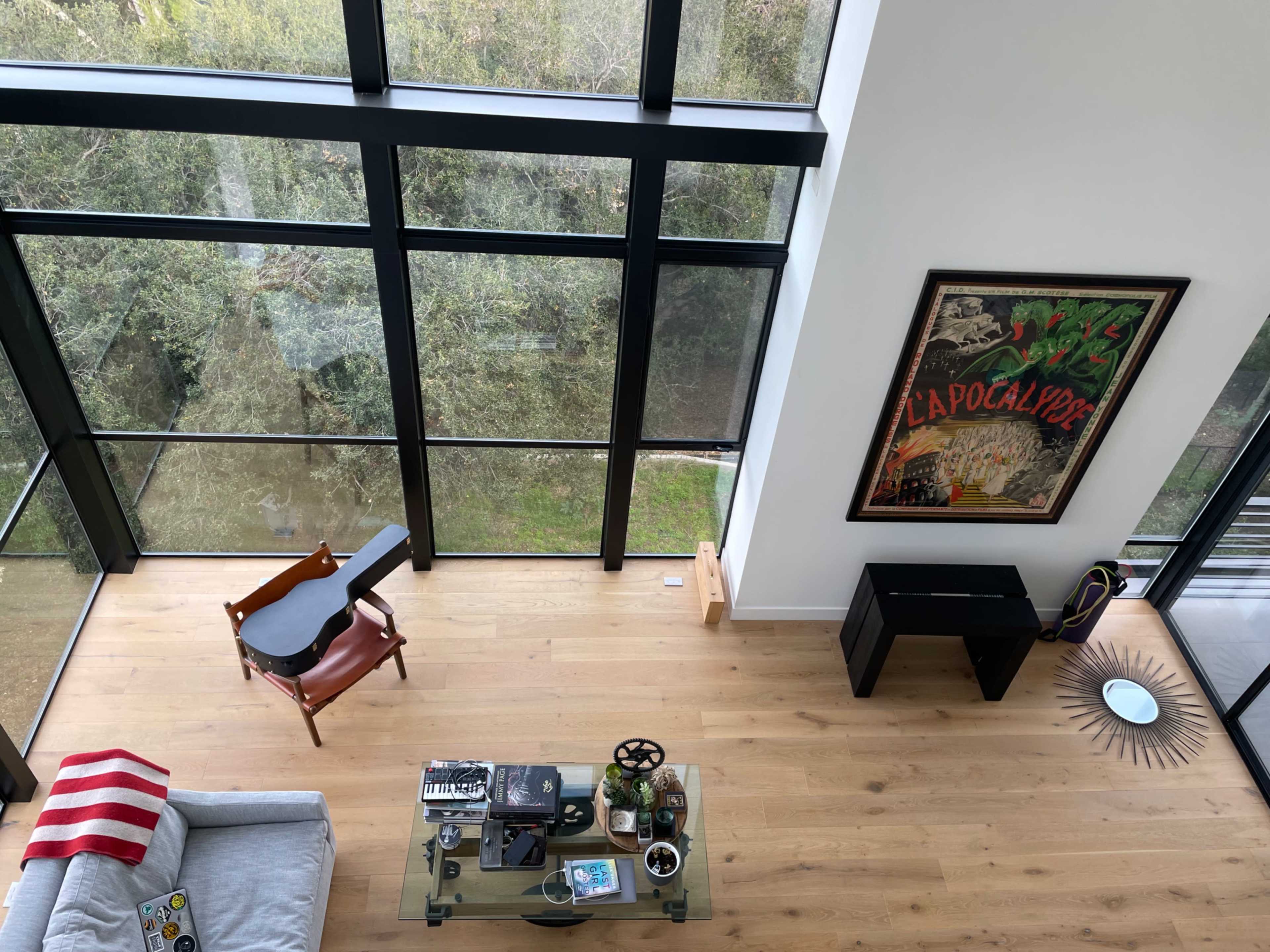 The image shows a modern living room with large windows, wooden flooring, a glass coffee table, a guitar, and a contemporary artwork on the wall.