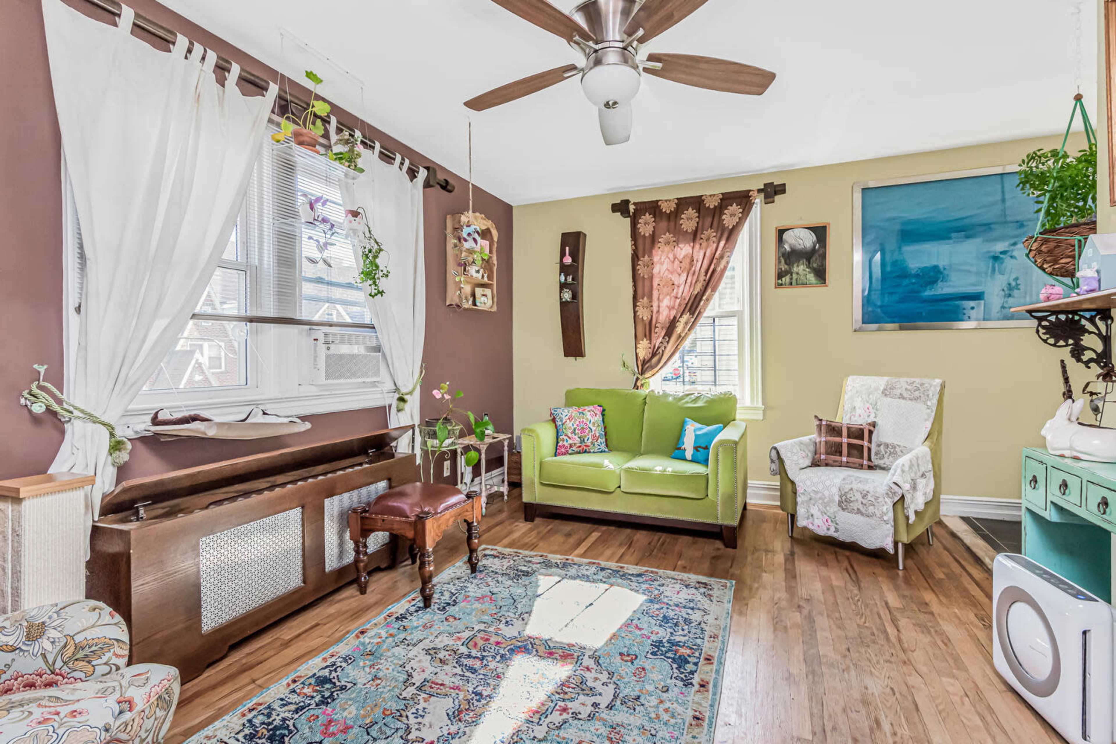 The image shows a cozy living room featuring a green sofa, a piano, and decorative plants, with a wooden floor and large windows allowing natural light.