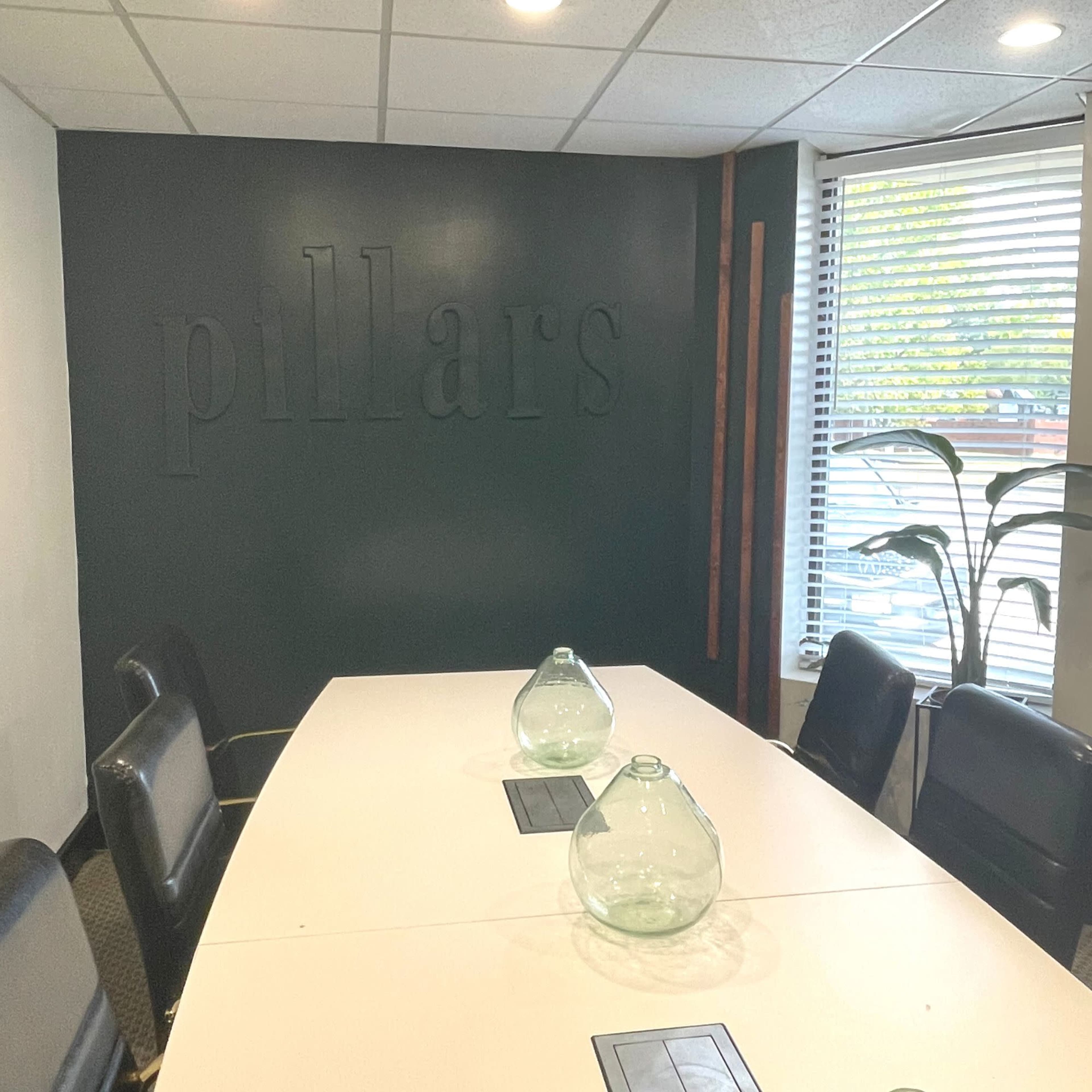 A conference room with a large white table, black chairs, and a green wall featuring the word "pillars" in raised letters.