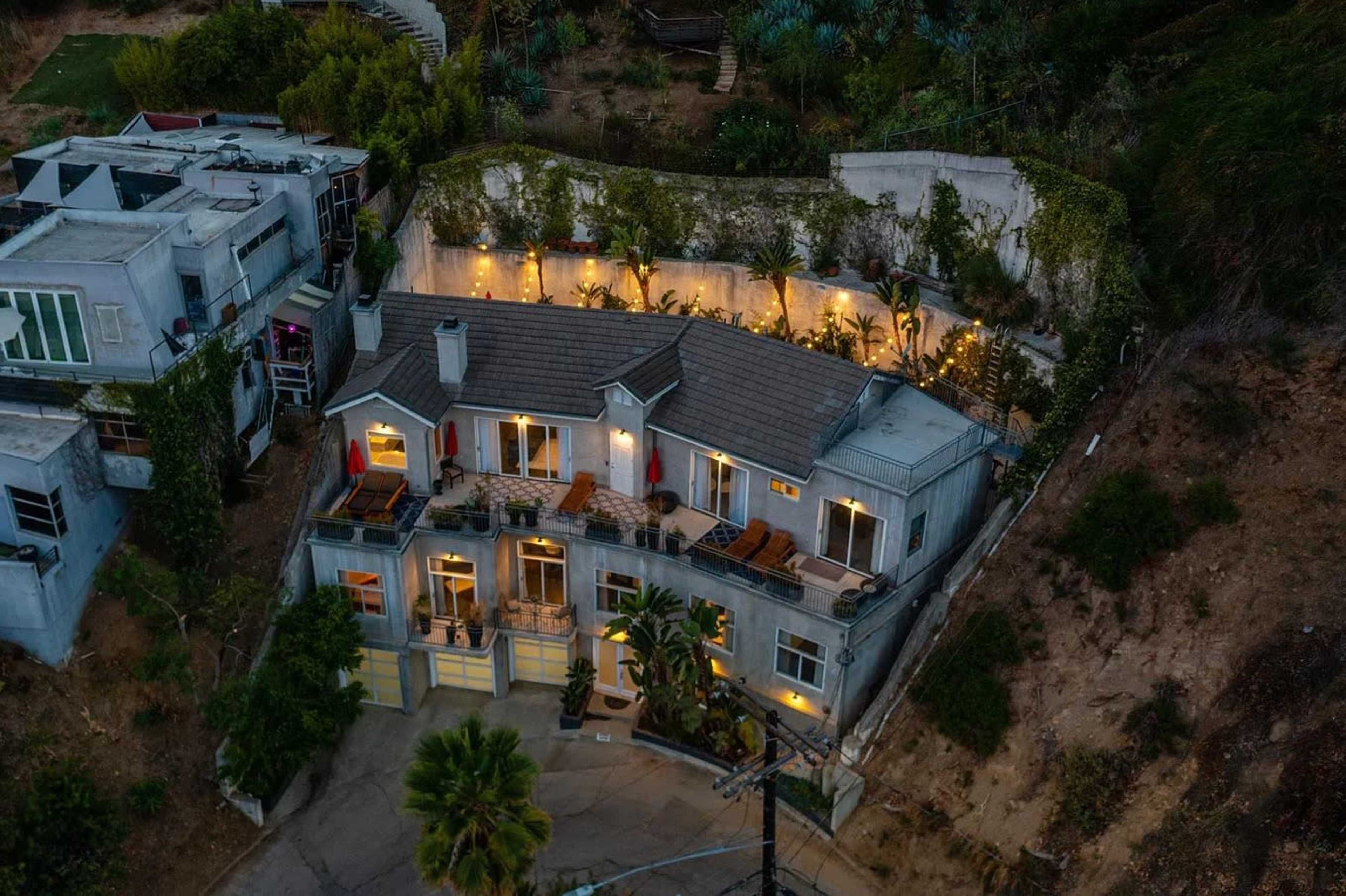A modern house situated on a hillside, surrounded by greenery and featuring outdoor lighting on its terraces.