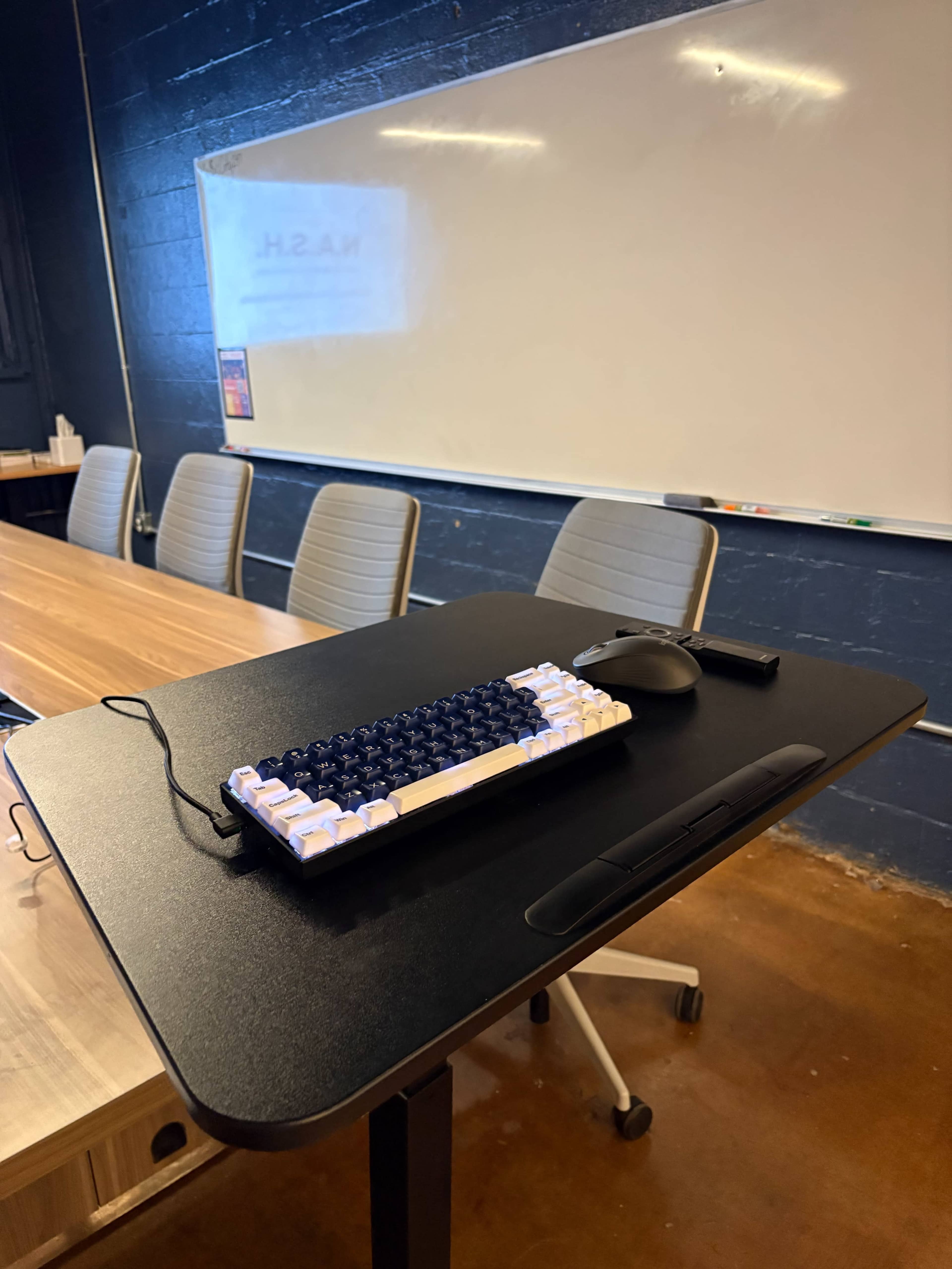 A black table with a mechanical keyboard and a mouse sits in front of a whiteboard in a conference room.