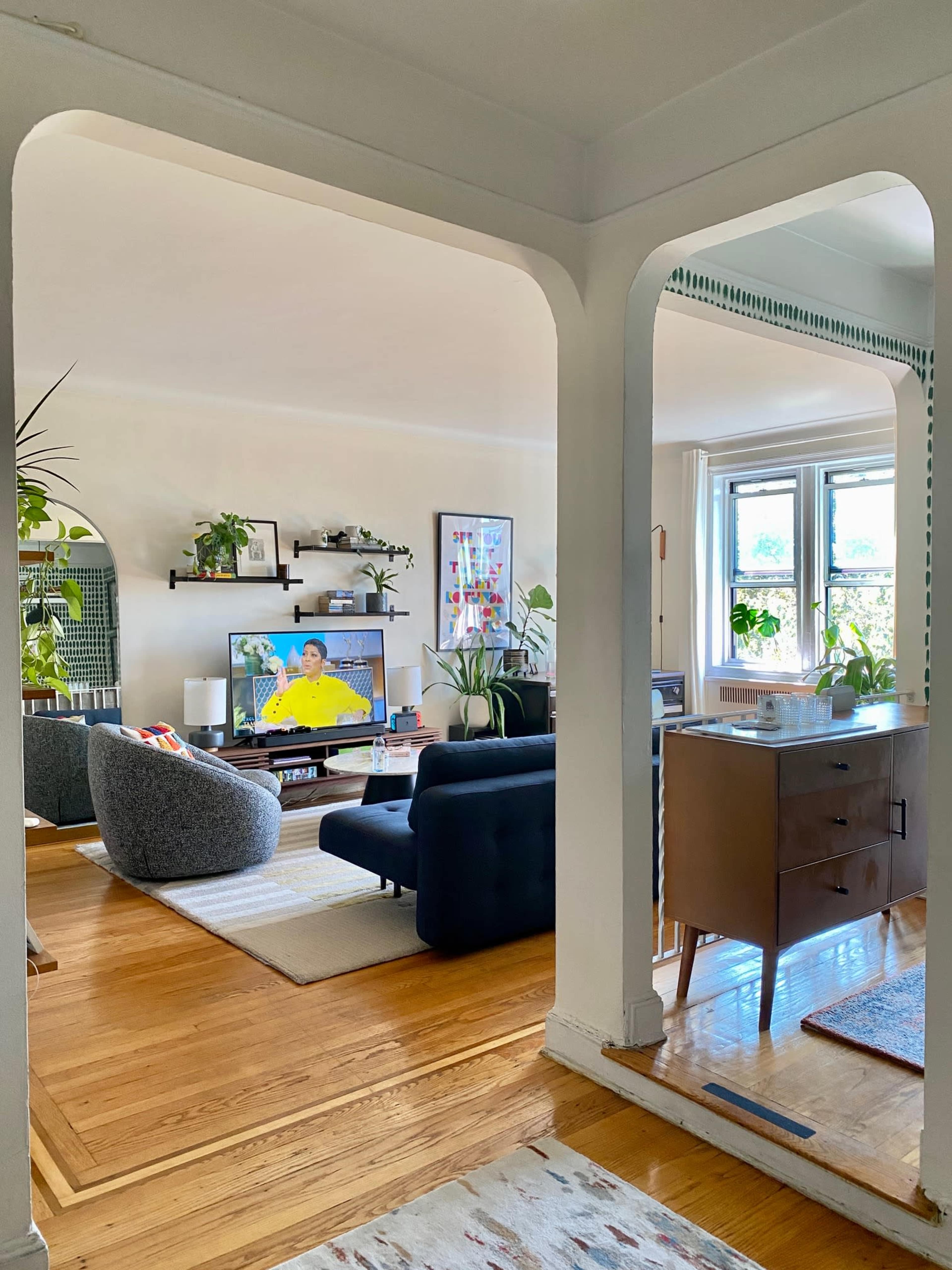 A bright living room features a blue sofa and a gray armchair, with a television displaying a program, framed by an archway and decorated with plants.