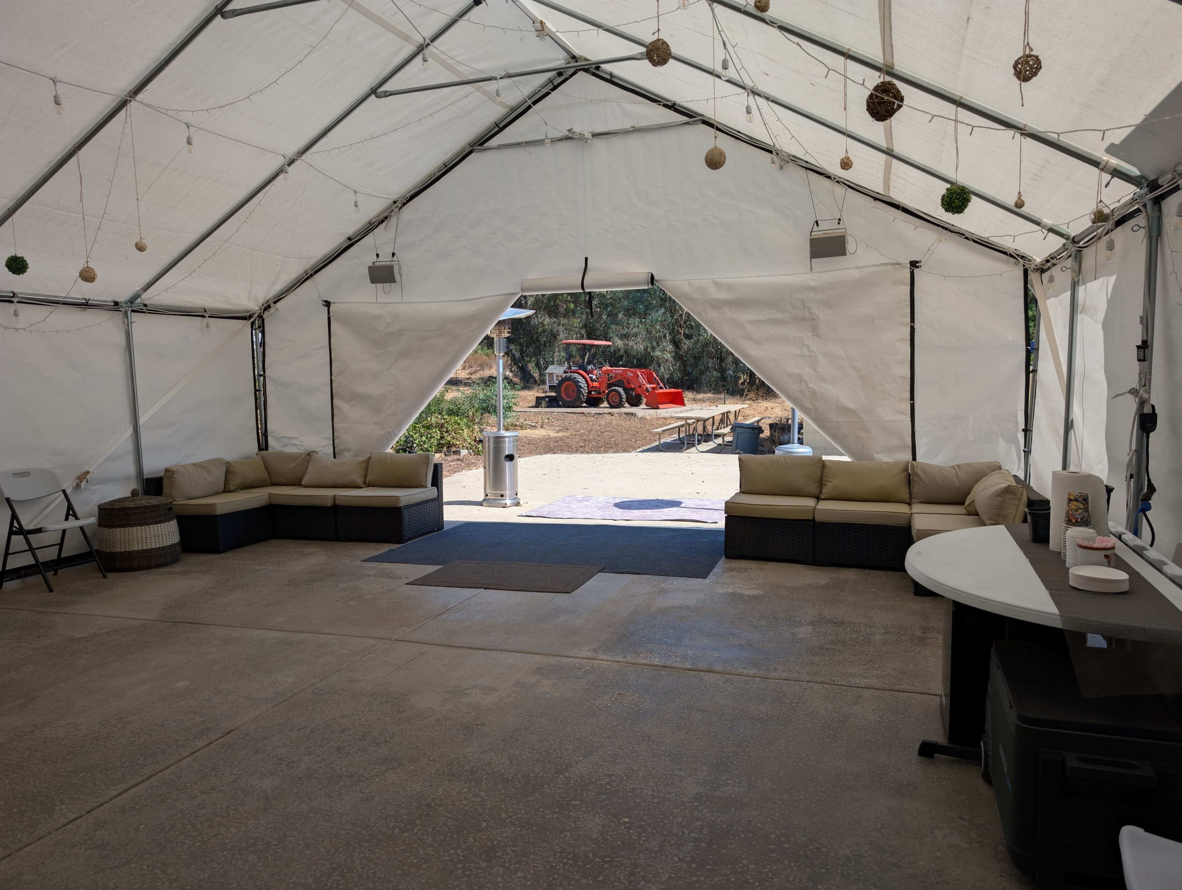 A spacious tent is set up with beige couches and a table, opening to an outdoor area where a red tractor is visible.