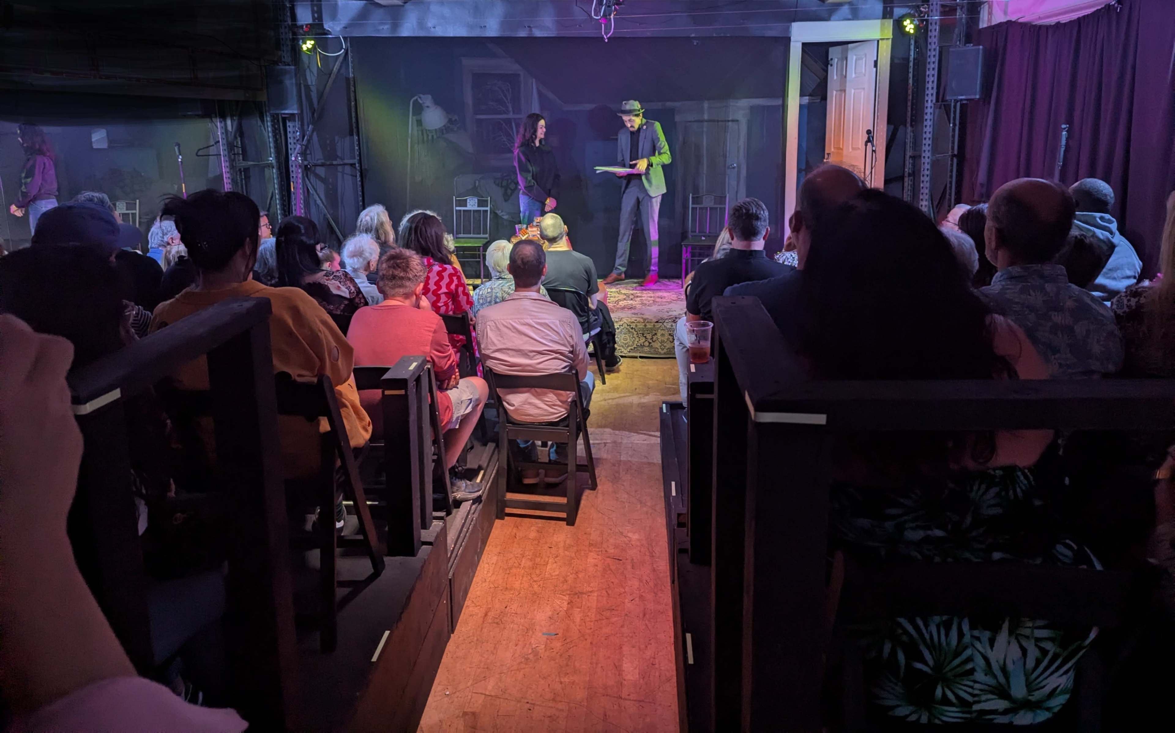 A crowd of seated audience members watches a performance on stage in a dimly lit venue.