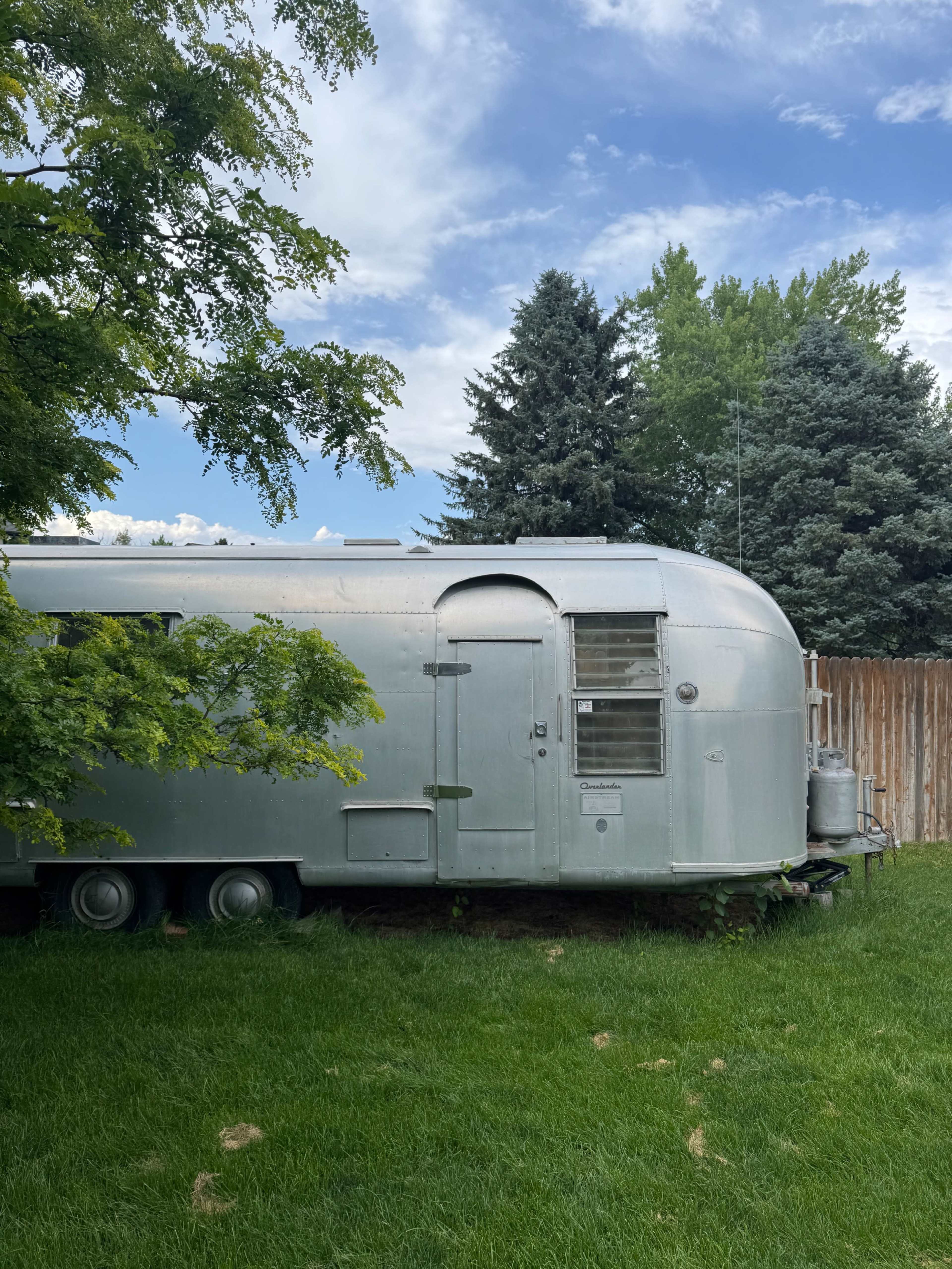 An Airstream trailer is parked on grass beside a wooden fence and surrounded by trees.