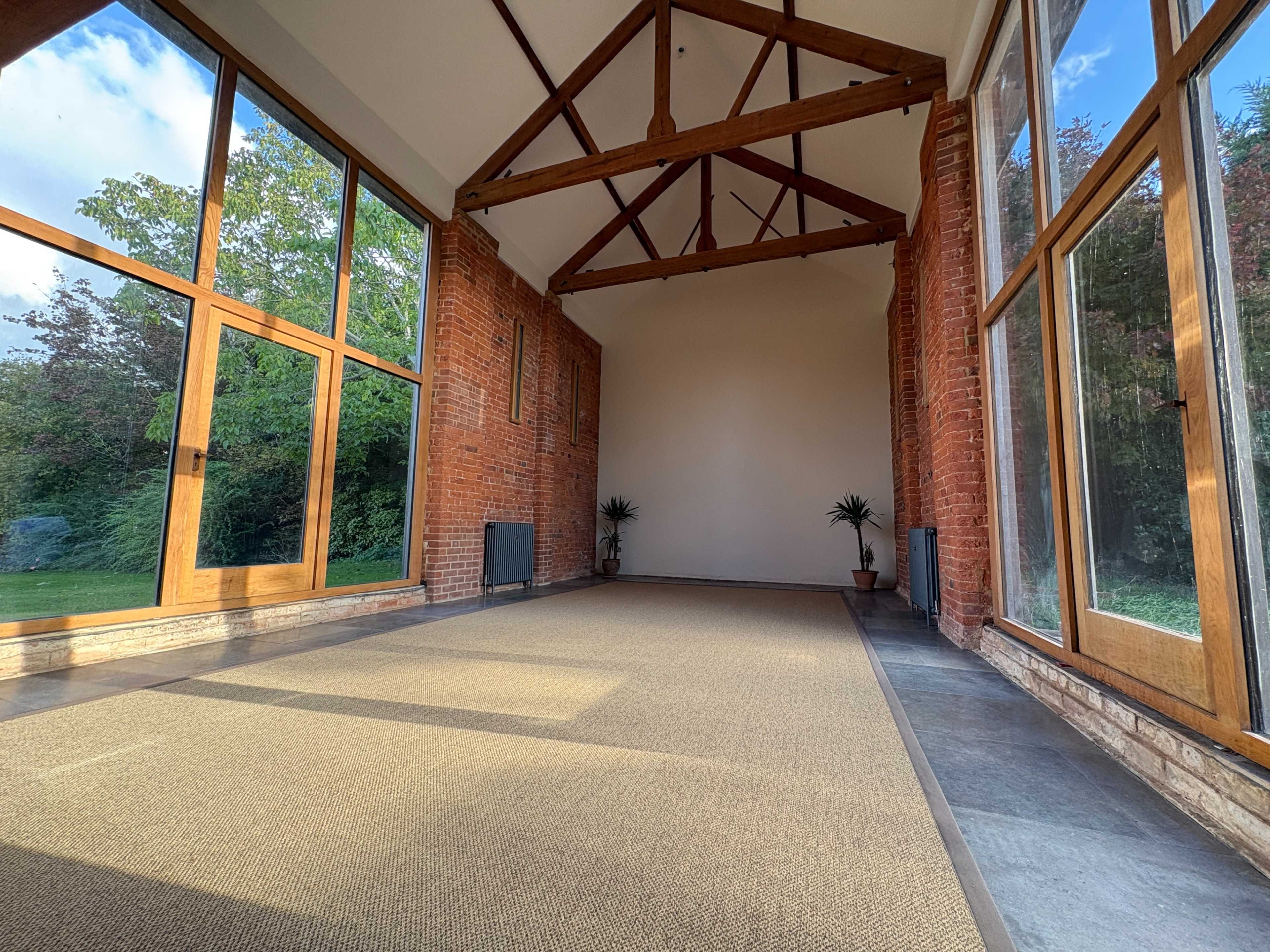 The image shows an interior space with large windows, exposed wooden beams, and a carpeted floor, surrounded by greenery outside.