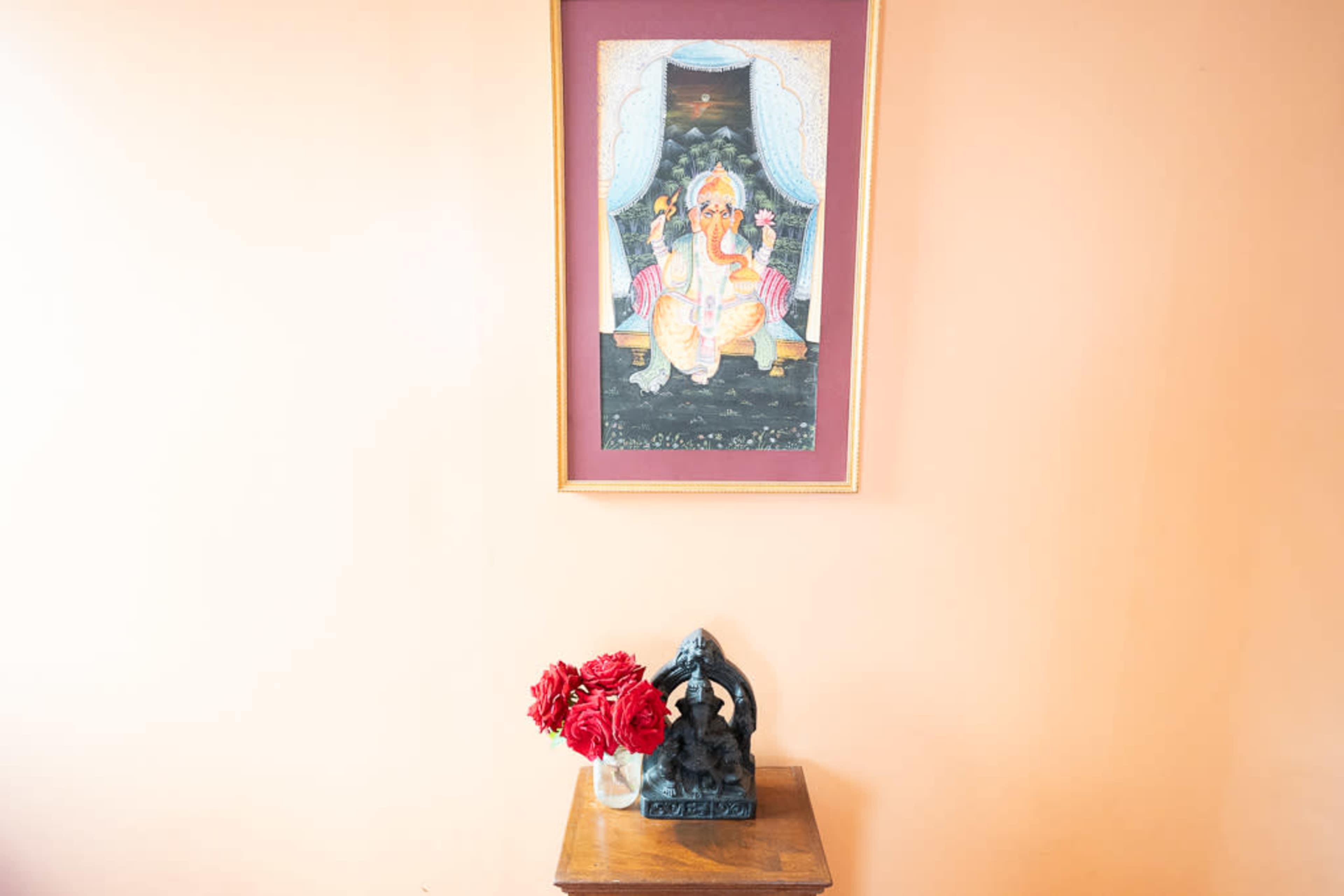 The image features a framed painting of a deity on a peach-colored wall, accompanied by a black statue and a vase of red flowers on a wooden table below.