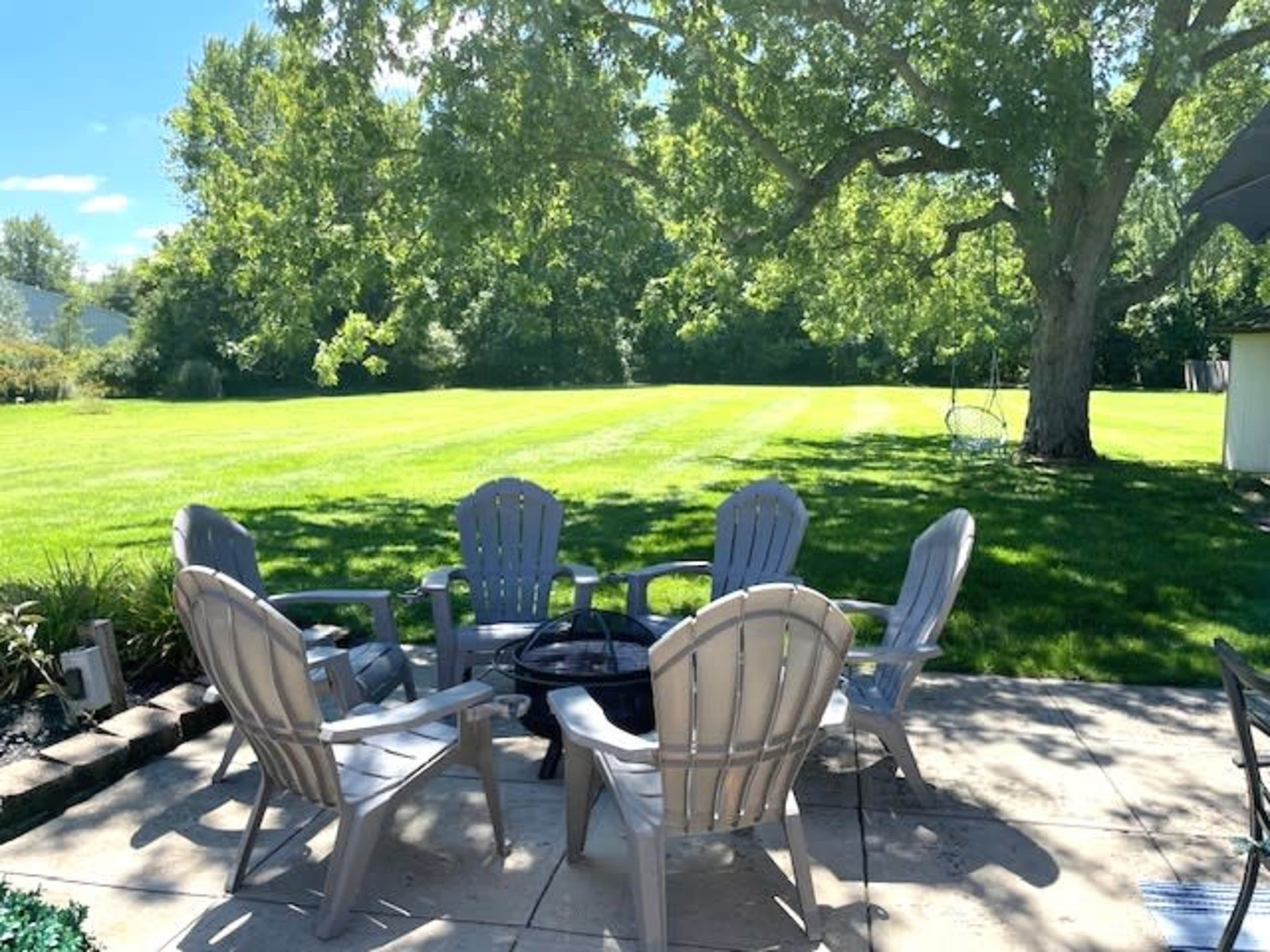 A patio area features six gray chairs arranged around a fire pit, overlooking a large grassy field and shaded by trees.