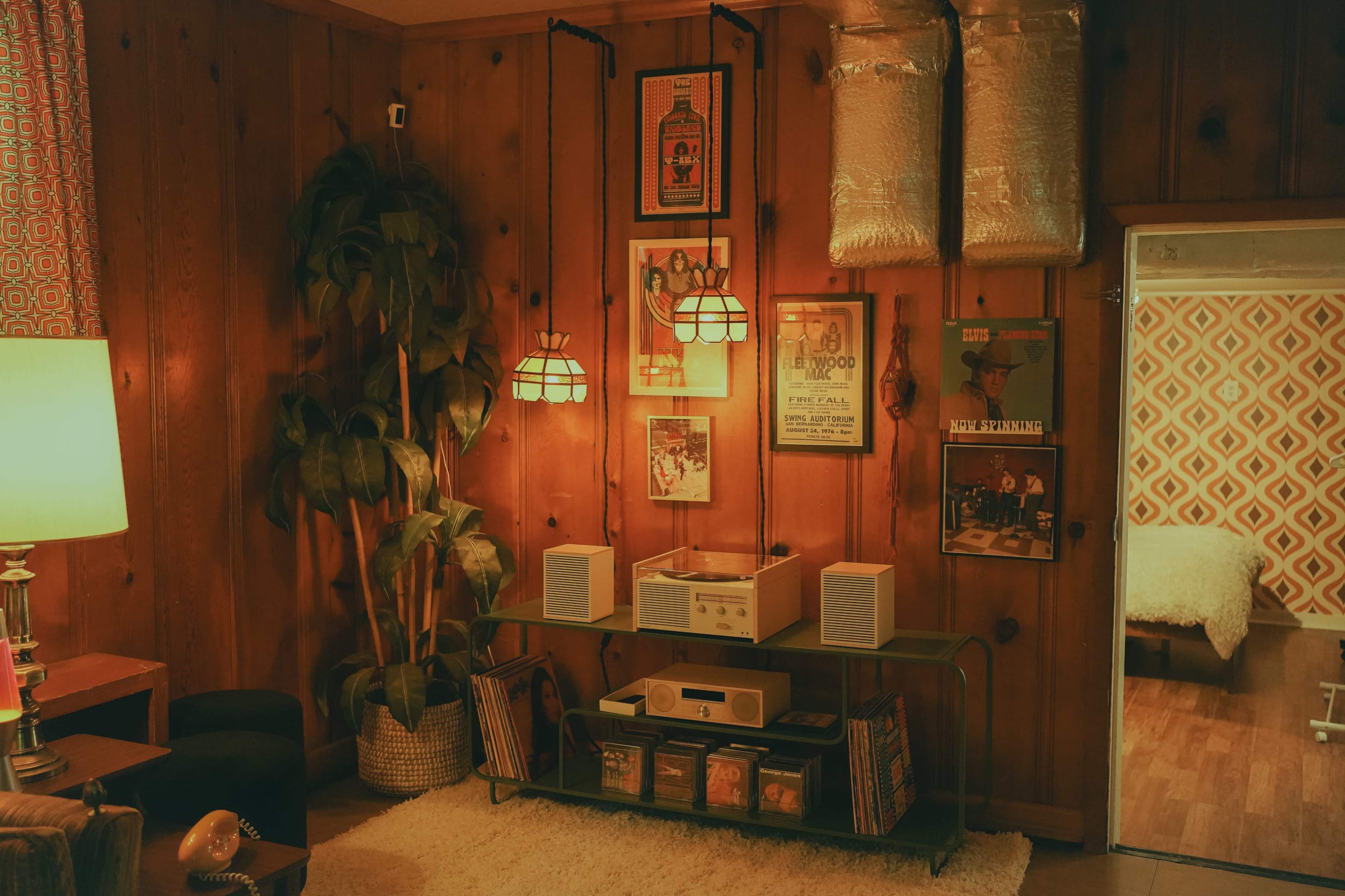 The scene depicts a vintage-style living room with wooden walls, hanging pendant lights, a potted plant, a record player, and a collection of vinyl records on display.