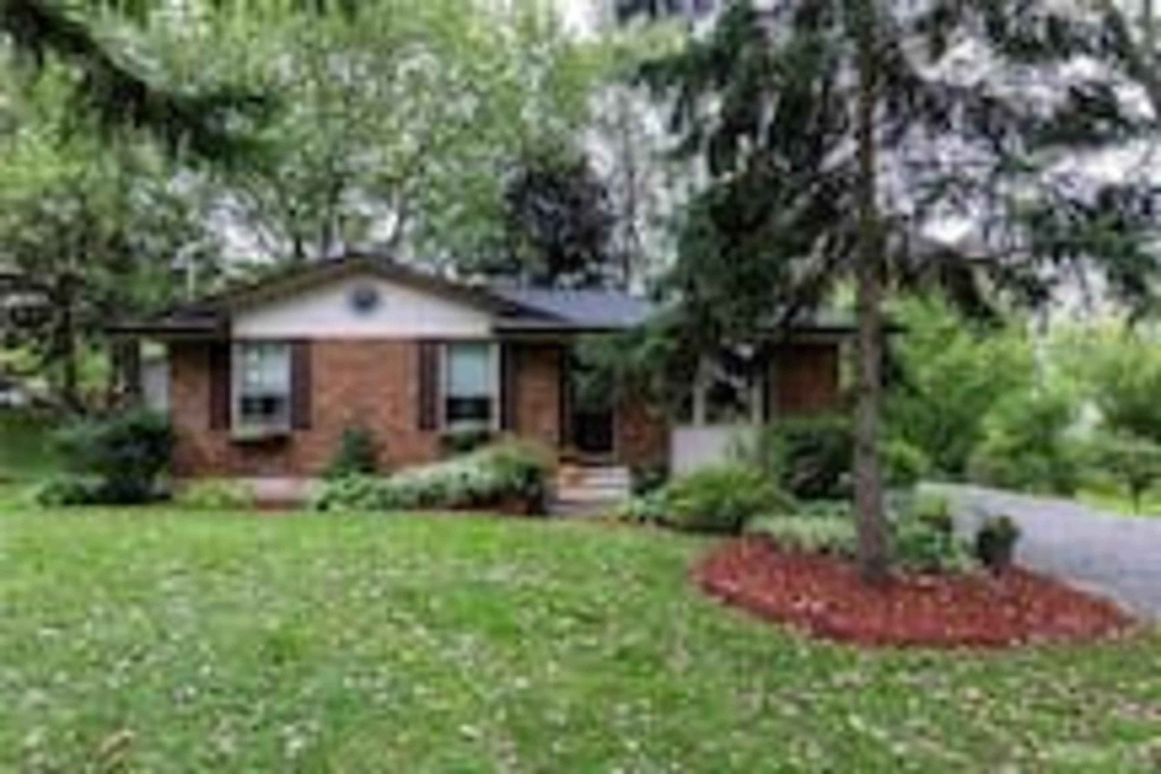 A single-story brick house is surrounded by a lush green lawn and landscaped gardens.