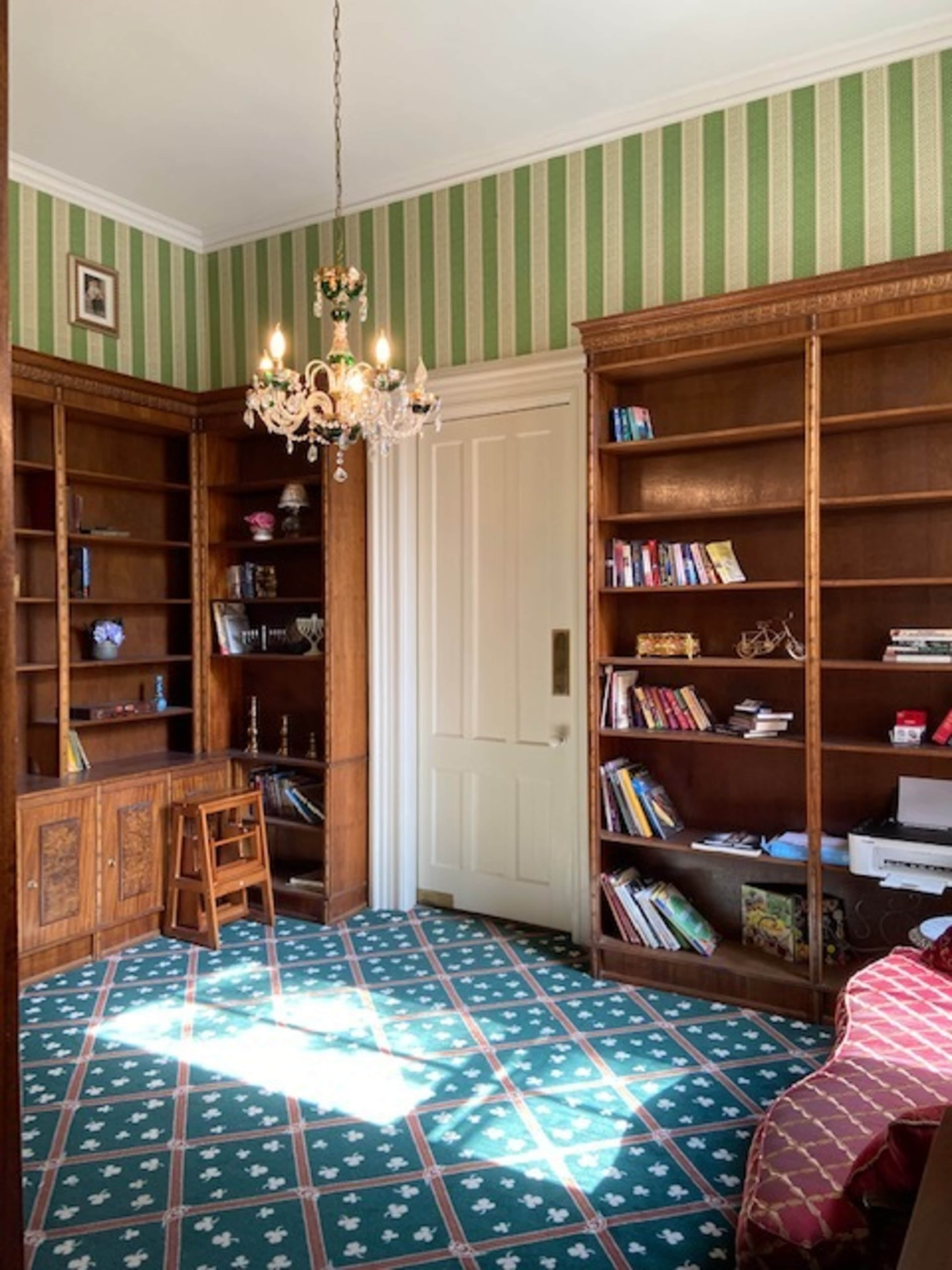 A room with wooden bookshelves filled with books, a small desk and chair, a door, and a chandelier hanging from the ceiling, all set against a backdrop of green striped wallpaper and a patterned carpet.