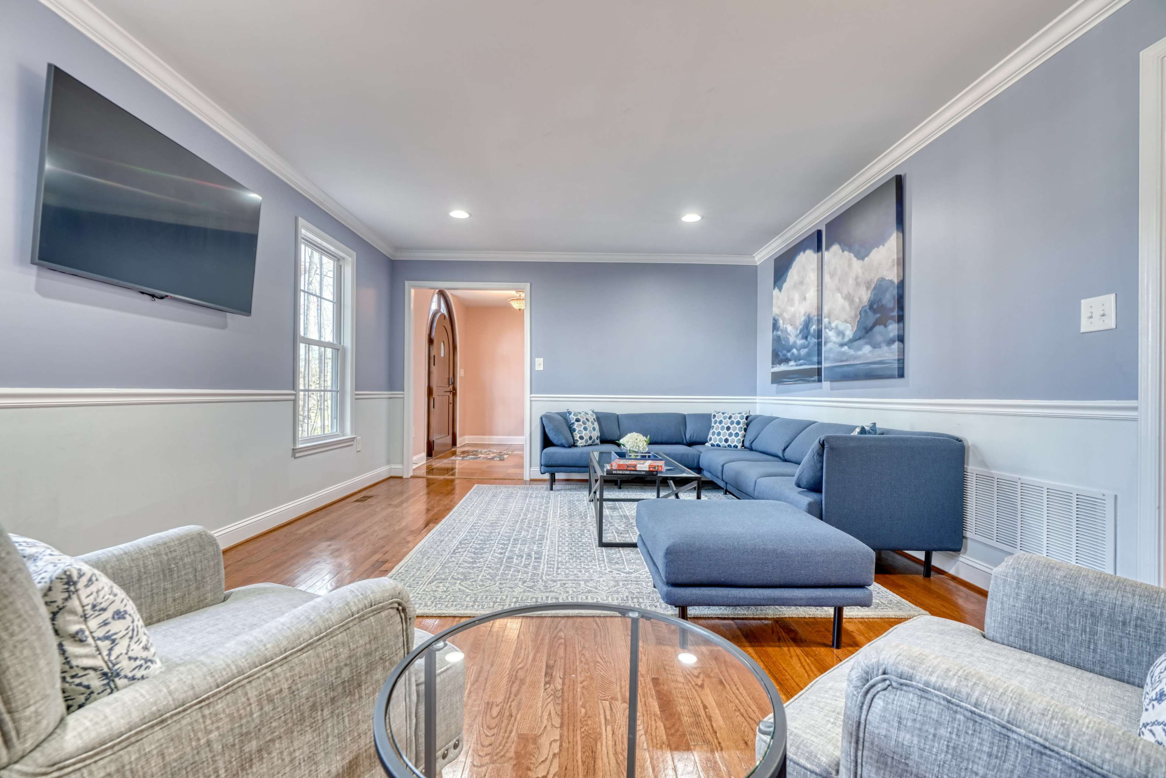 The living room features a gray sectional sofa, two armchairs, a glass coffee table, and a mounted television along with a light blue wall and hardwood flooring.