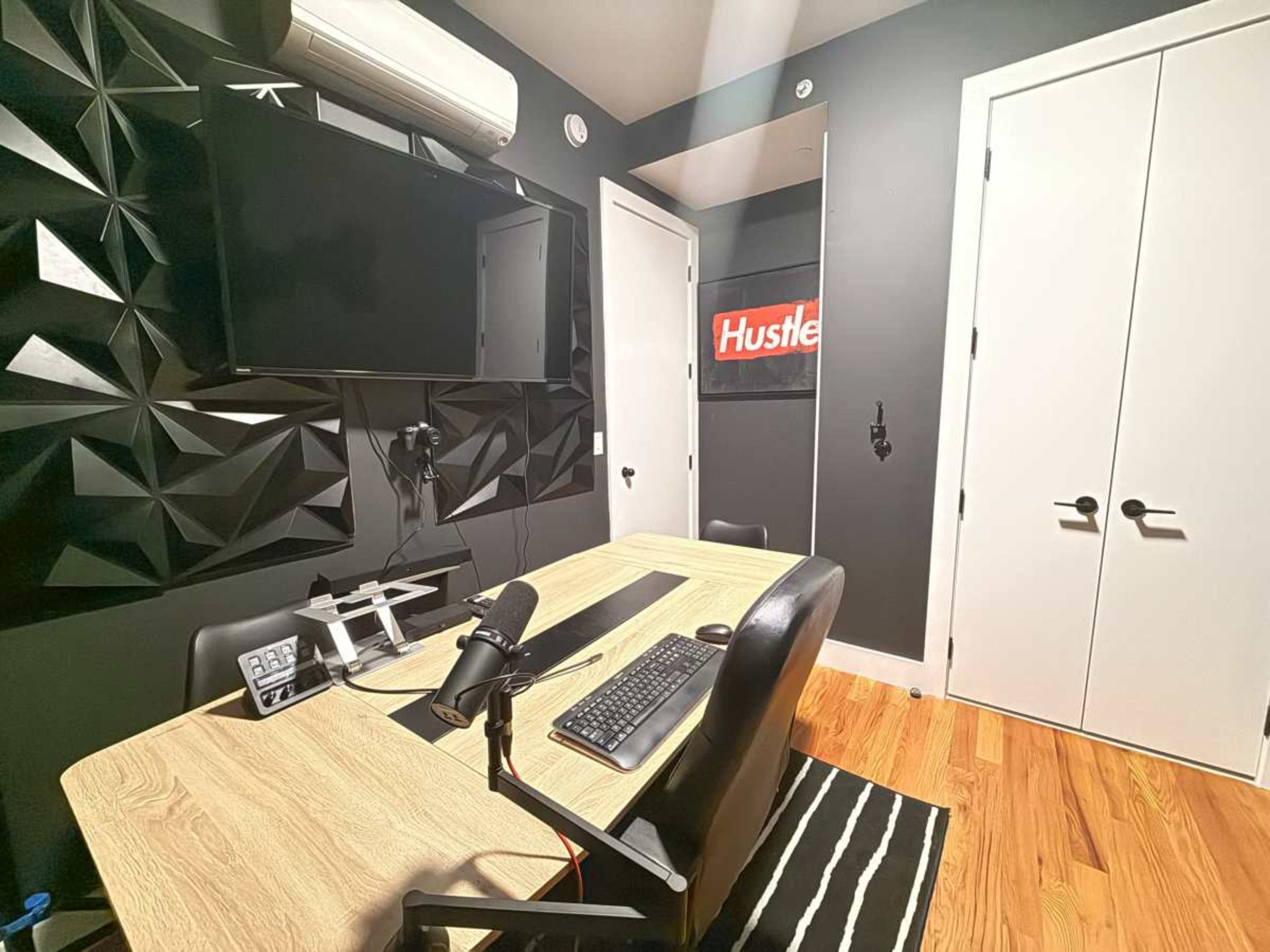 The image shows a modern home office with a wooden desk, computer setup, and black geometric wall panels, featuring a red "Hustle" sign on the wall.