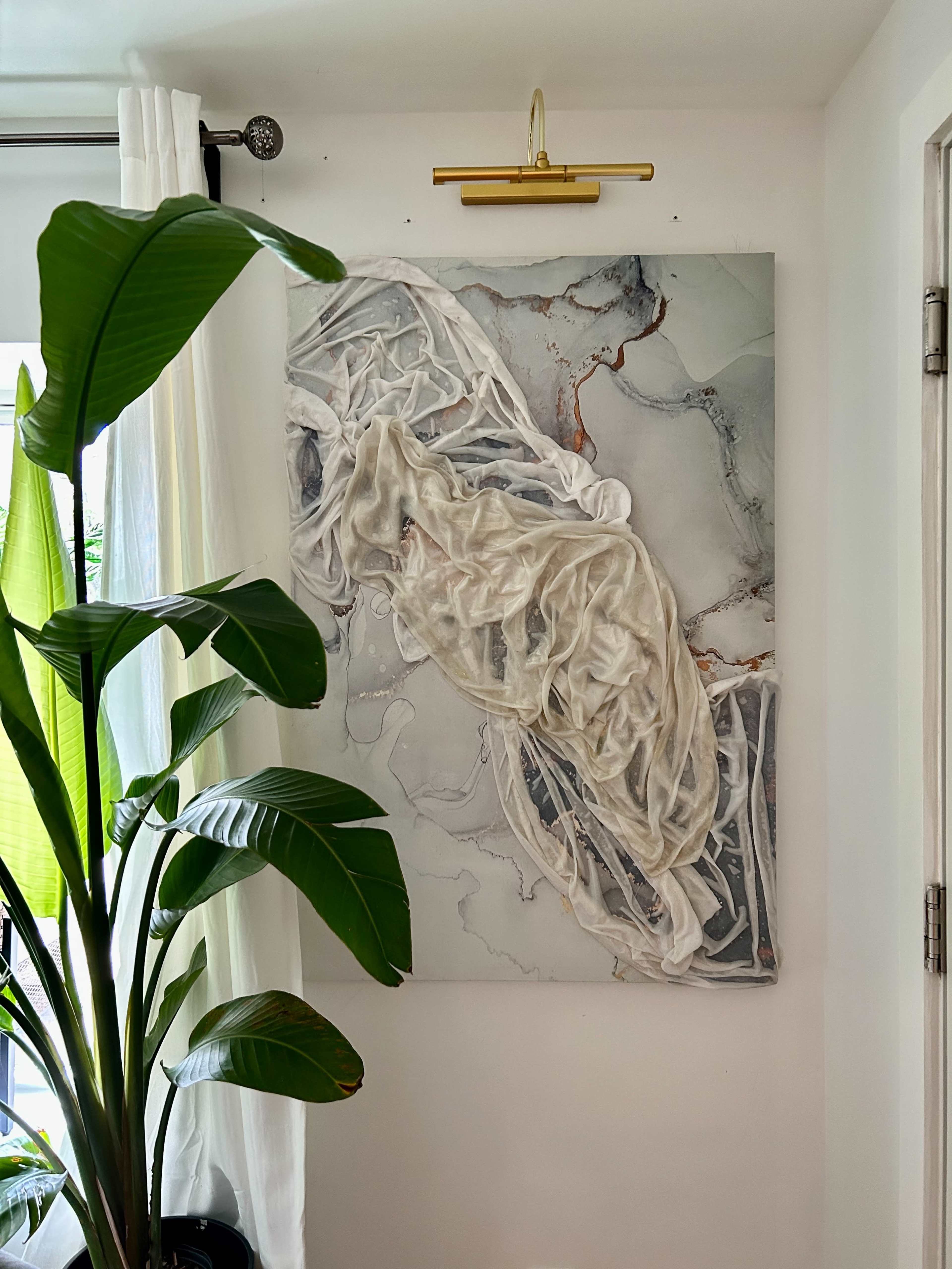 A large artwork featuring draped white fabric against a light gray background, hung on a wall next to a tall green plant.