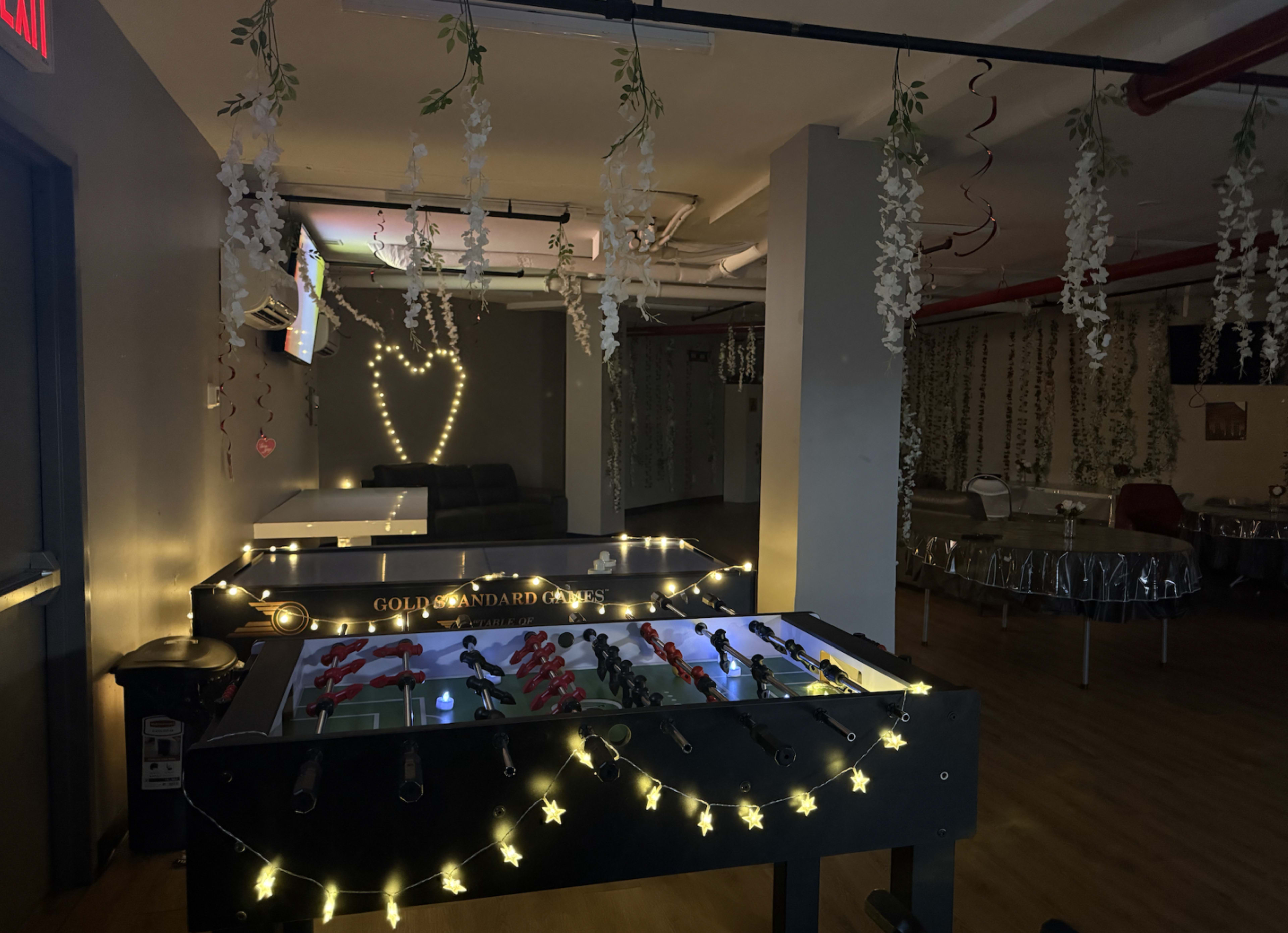 The image shows a dimly lit game room with foosball tables adorned with string lights and decorative vines hanging from the ceiling.