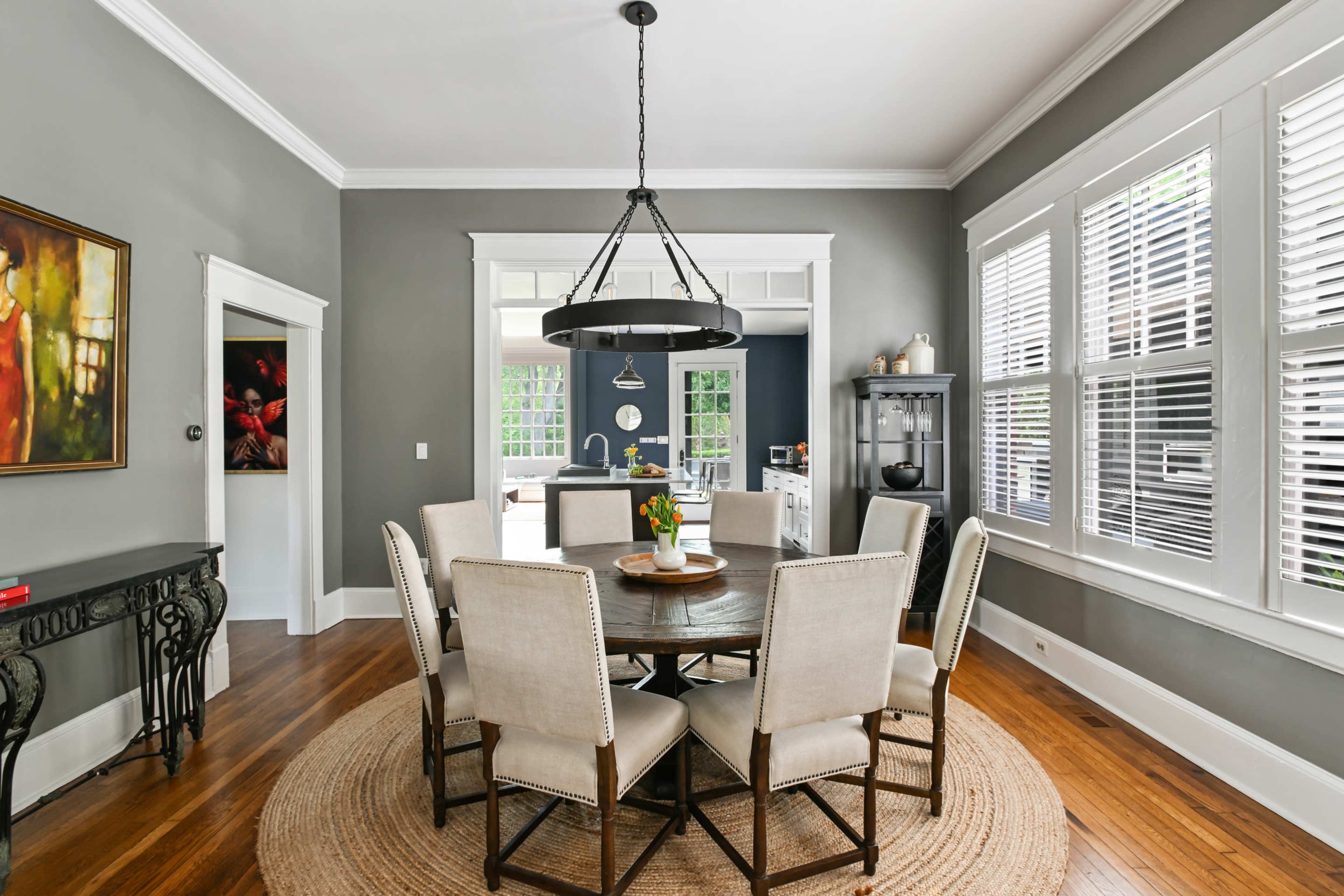 A round dining table with eight upholstered chairs is set in a well-lit room featuring gray walls and large windows.