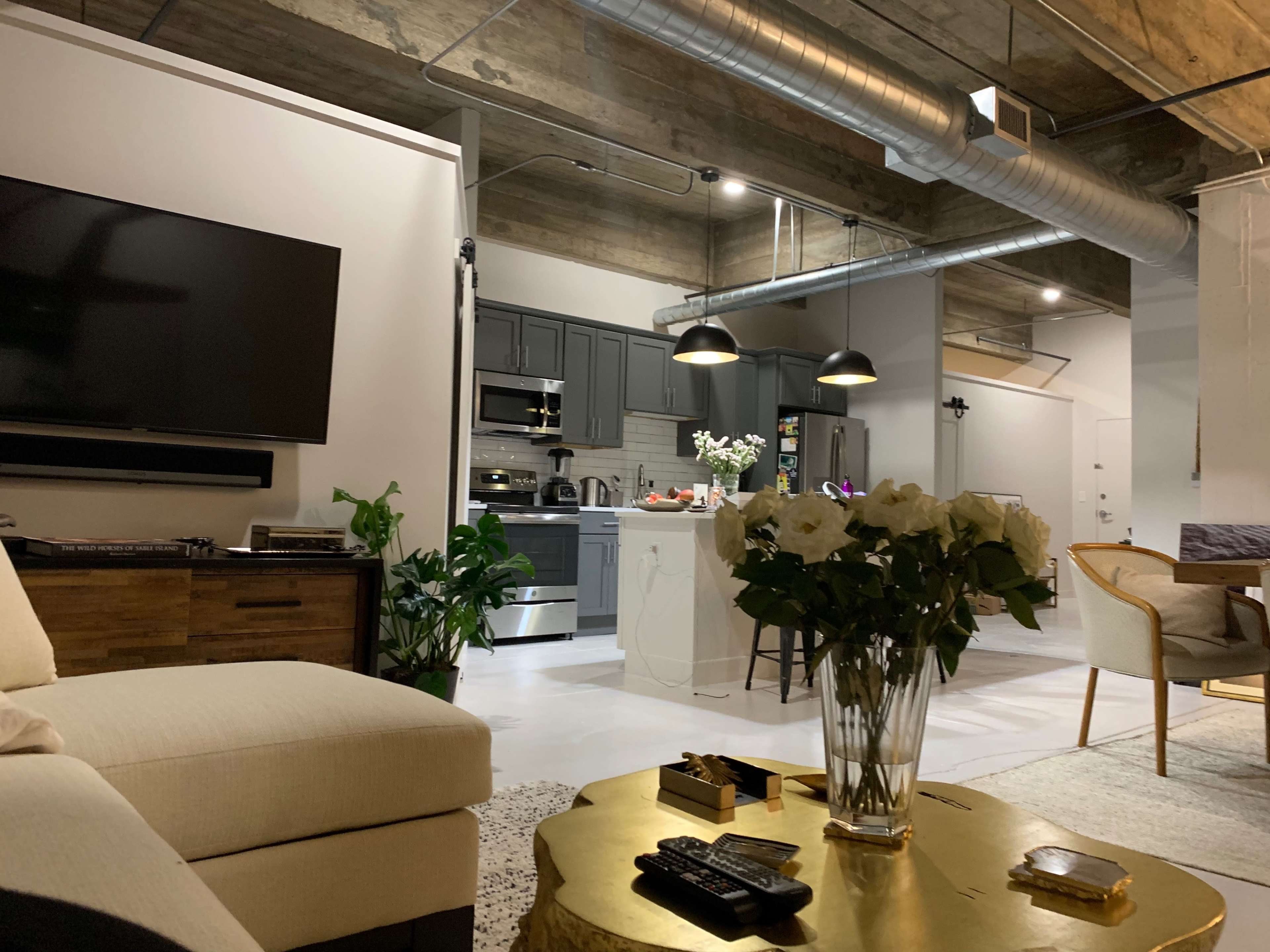 A modern, open-concept living space featuring a comfortable couch, a television mounted on the wall, a kitchen in the background, and a dining area with a decorative table.
