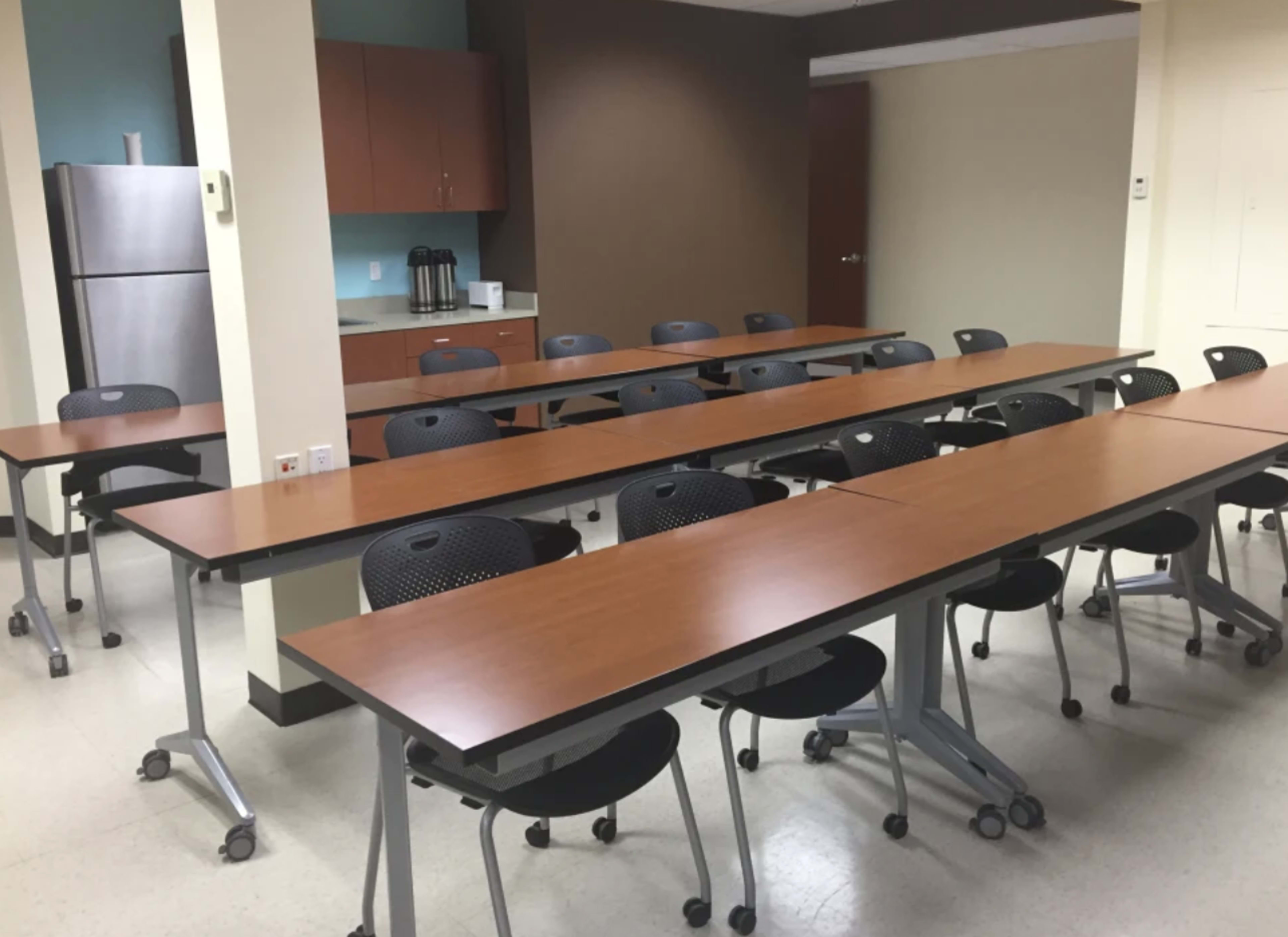 A conference room with multiple long tables arranged in rows and black chairs positioned around them, along with a kitchenette in the background.