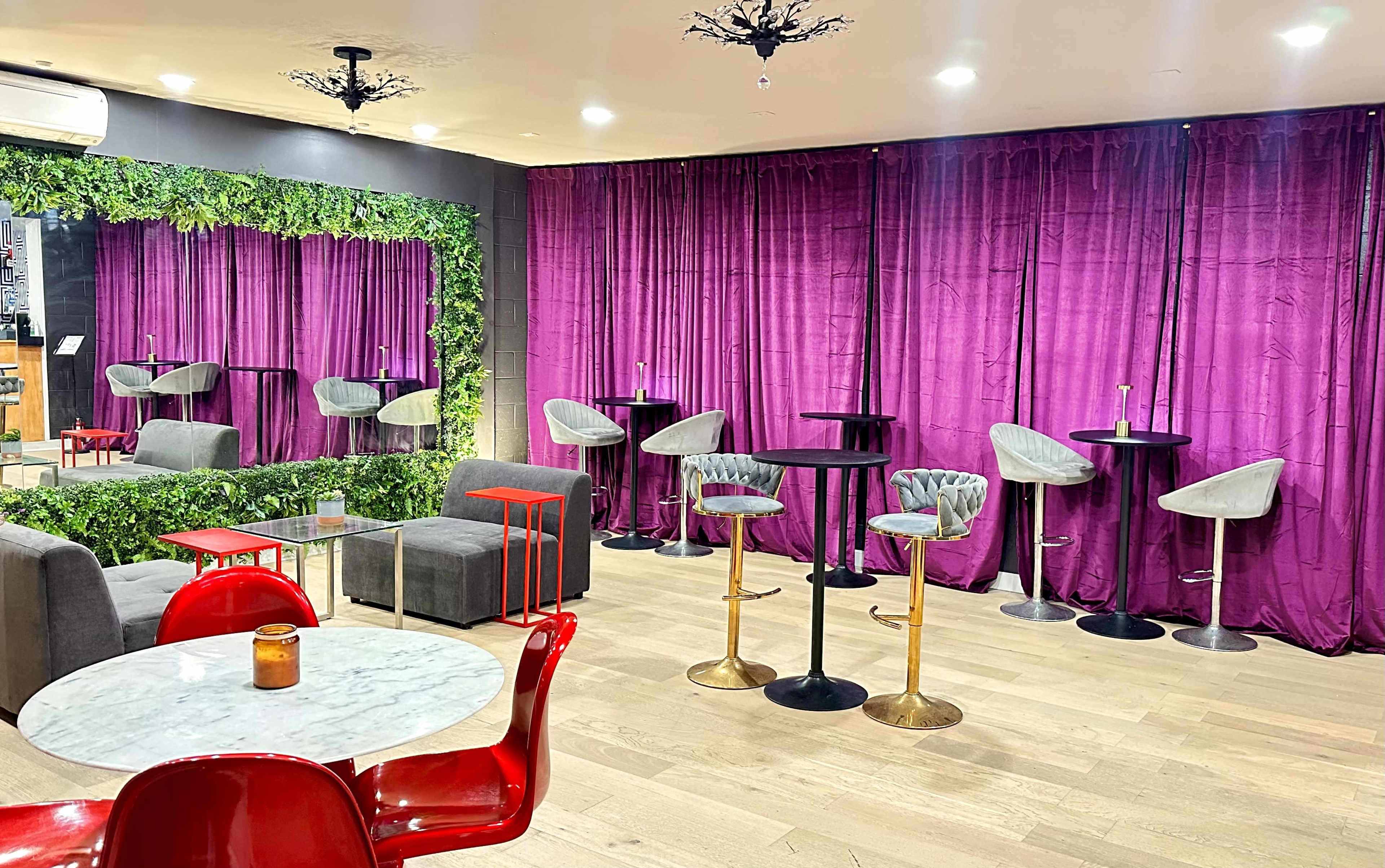 The image shows a stylish lounge area featuring purple curtains, green foliage, seating arrangements with gray chairs and stools, and a marble table with red and gold accents.