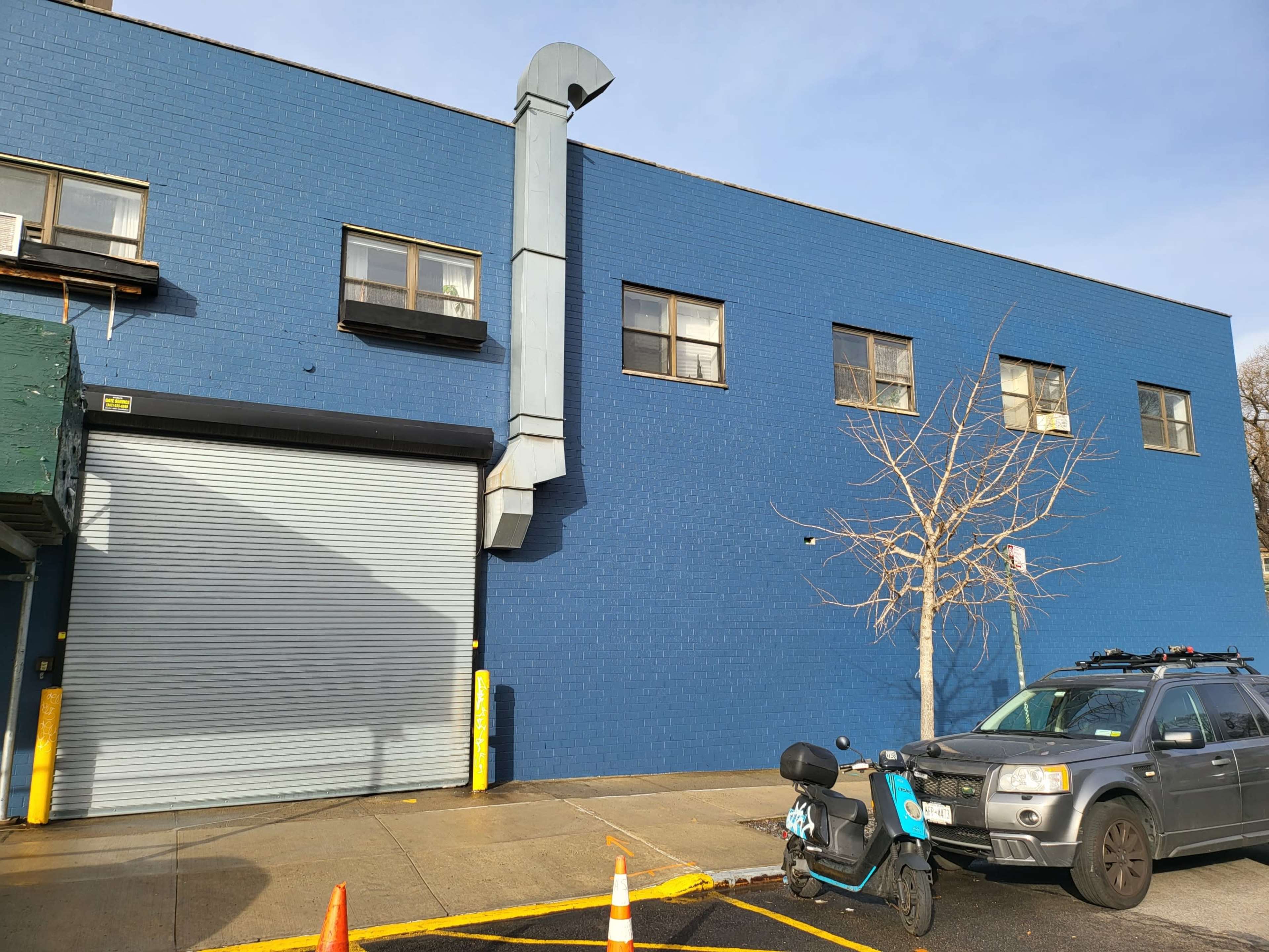 A blue brick building features a large garage door, a vertical ventilation pipe, and a parked police vehicle beside a tree.