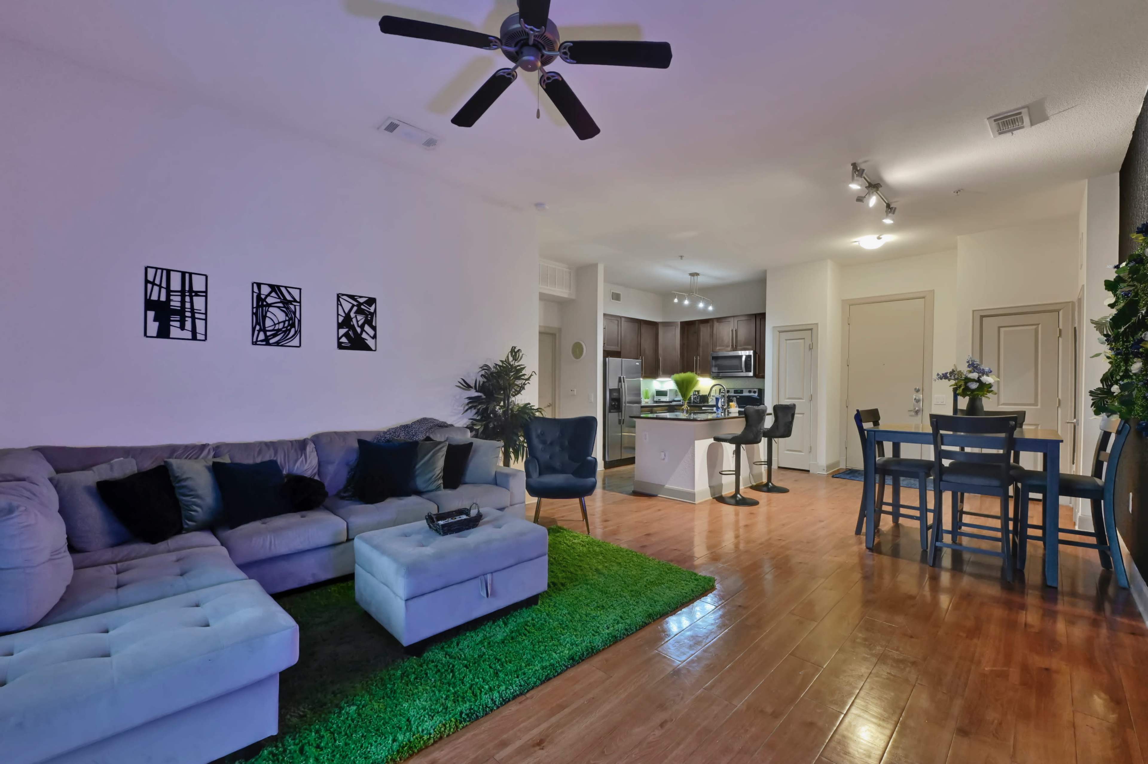 The image shows a modern living room with a gray sectional sofa, a green rug, and a view of an adjacent kitchen and dining area.