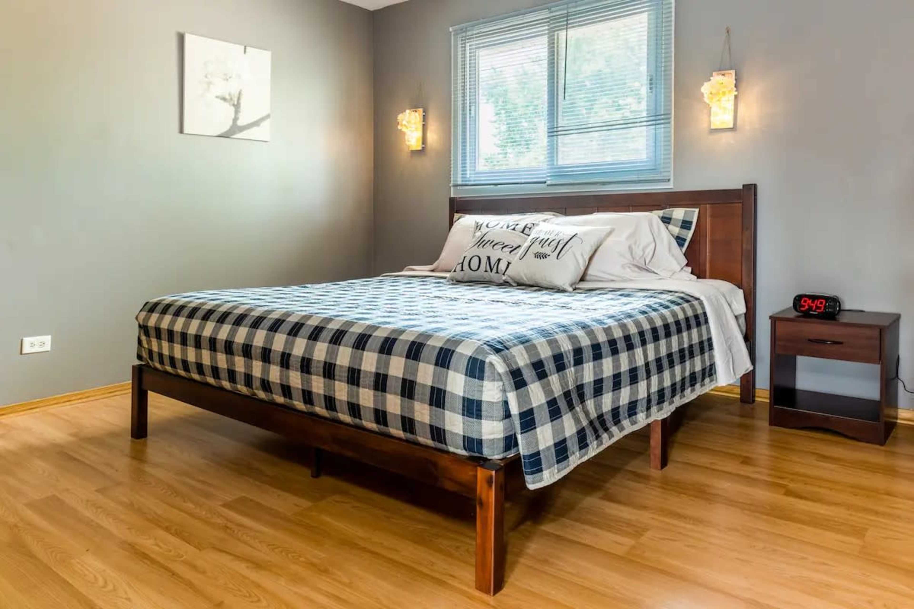 The image shows a neatly made bed with a plaid pattern in a light-colored bedroom featuring wooden flooring and a nightstand with a clock.