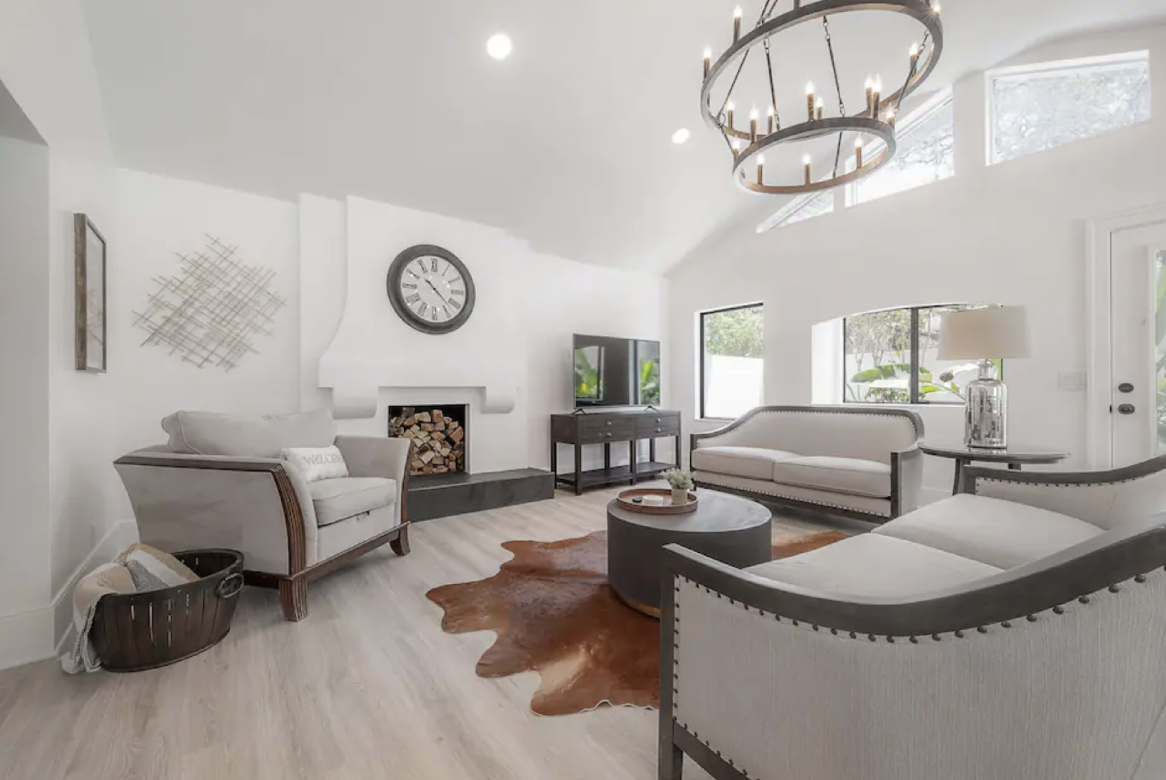 The living room features light-colored walls, a modern chandelier, a fireplace with stacked logs, and minimalist furnishings, including a circular coffee table and two sofas.