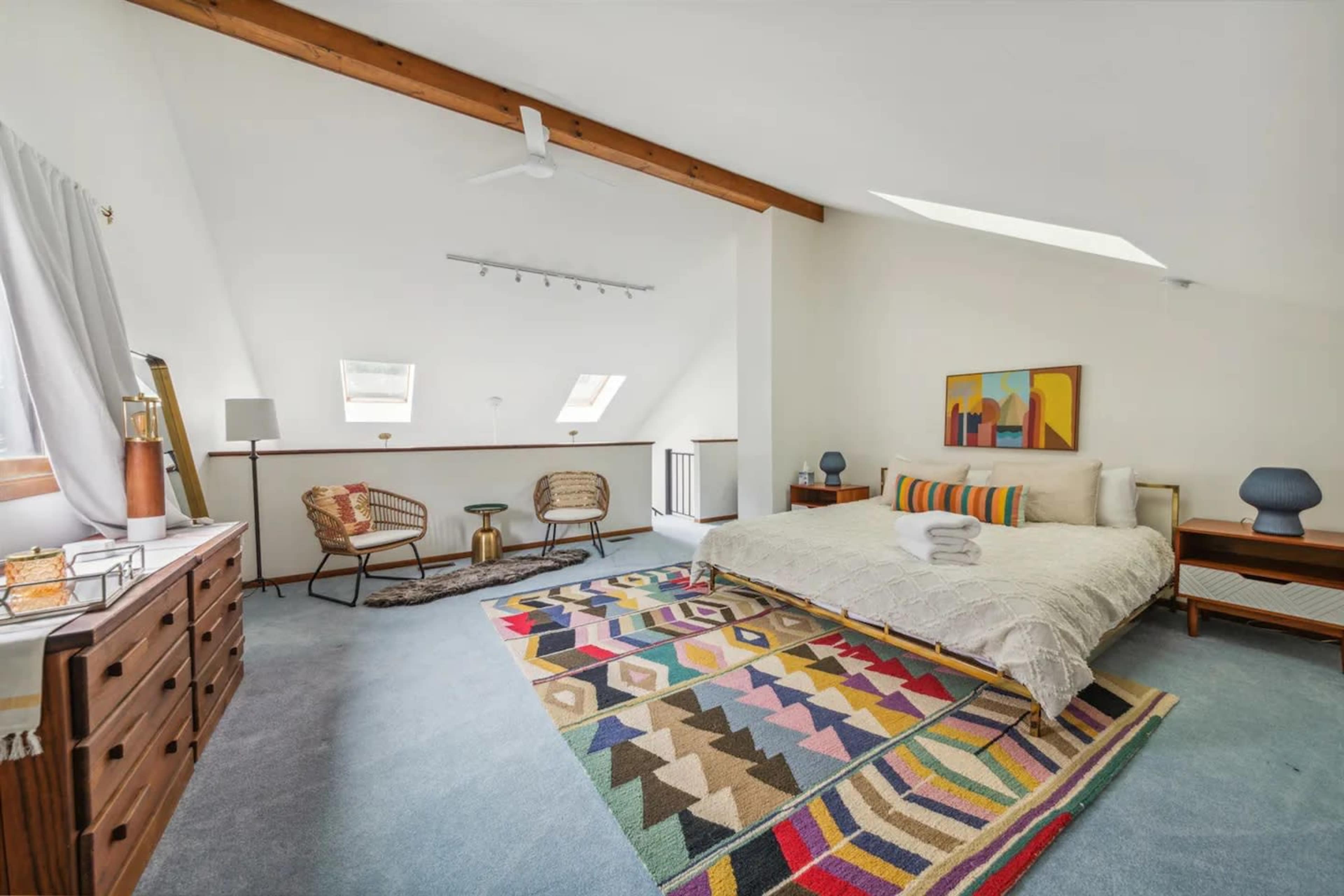 An attic bedroom featuring a large bed with a patterned rug, wooden furniture, and sloped ceilings with skylights.
