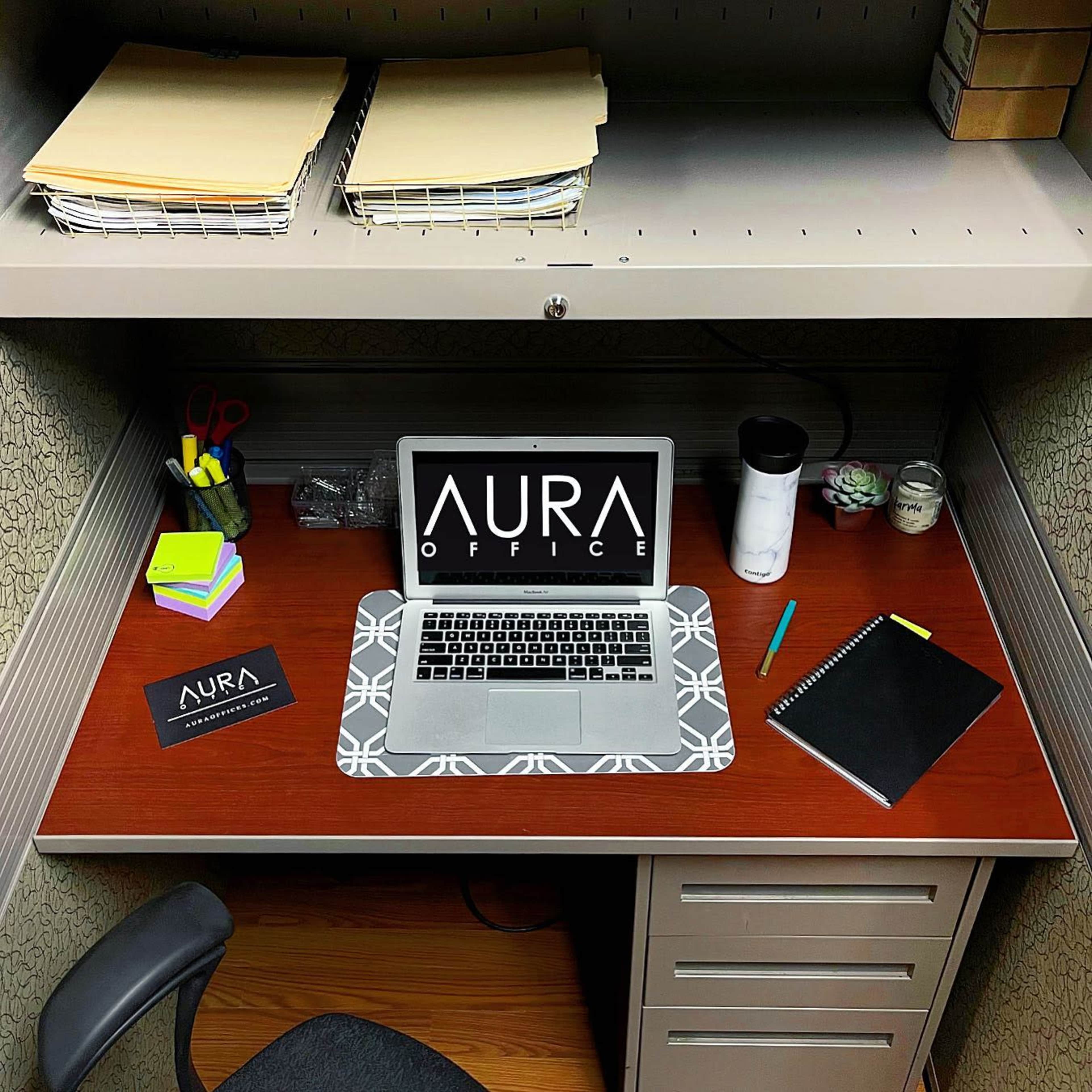 A neatly arranged office desk features a laptop, stationery supplies, a notebook, and a branded card, all set against a gray cubicle backdrop.