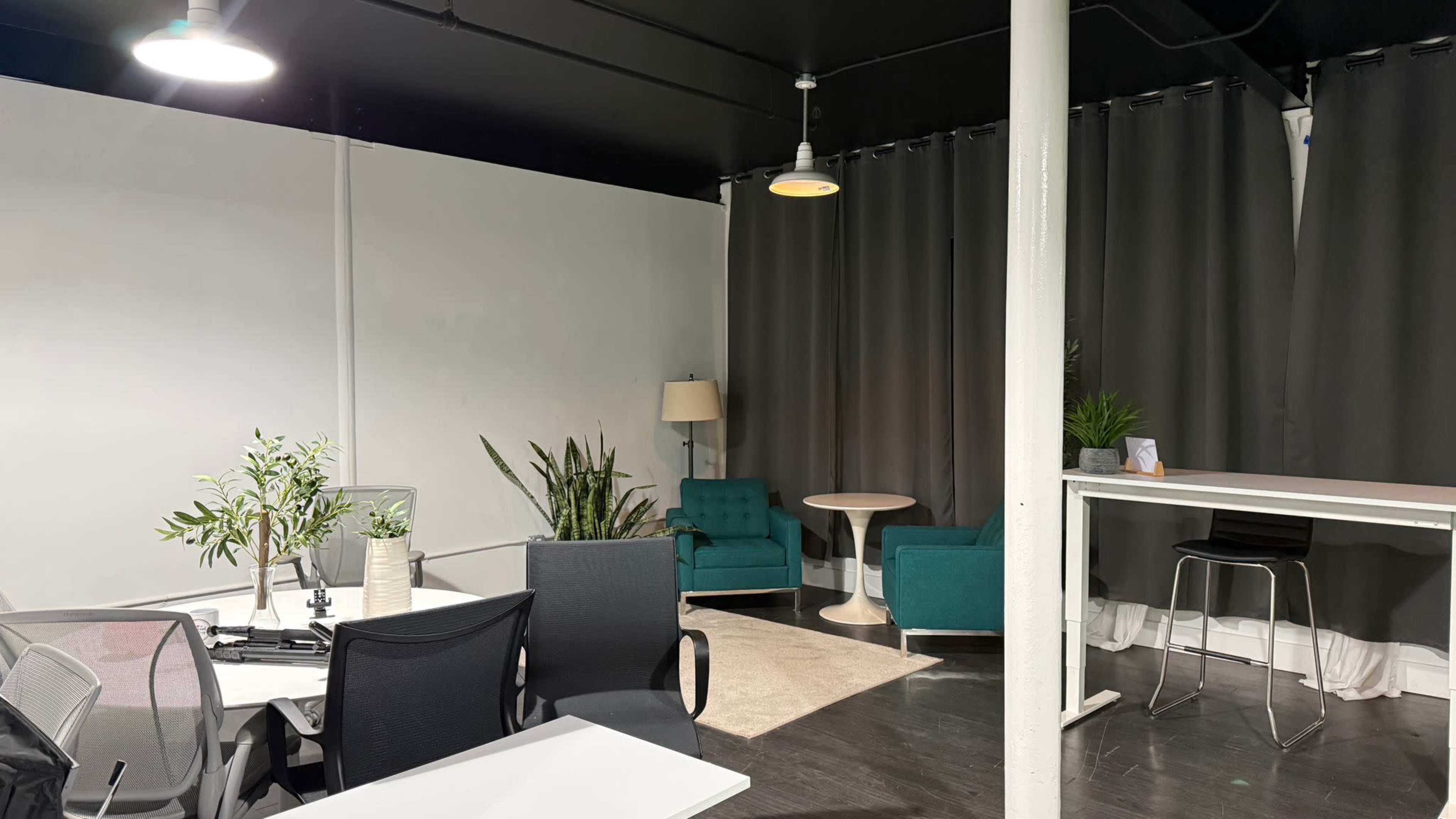 The image shows a modern office space with two green chairs, a small round table, various potted plants, and several workstations.