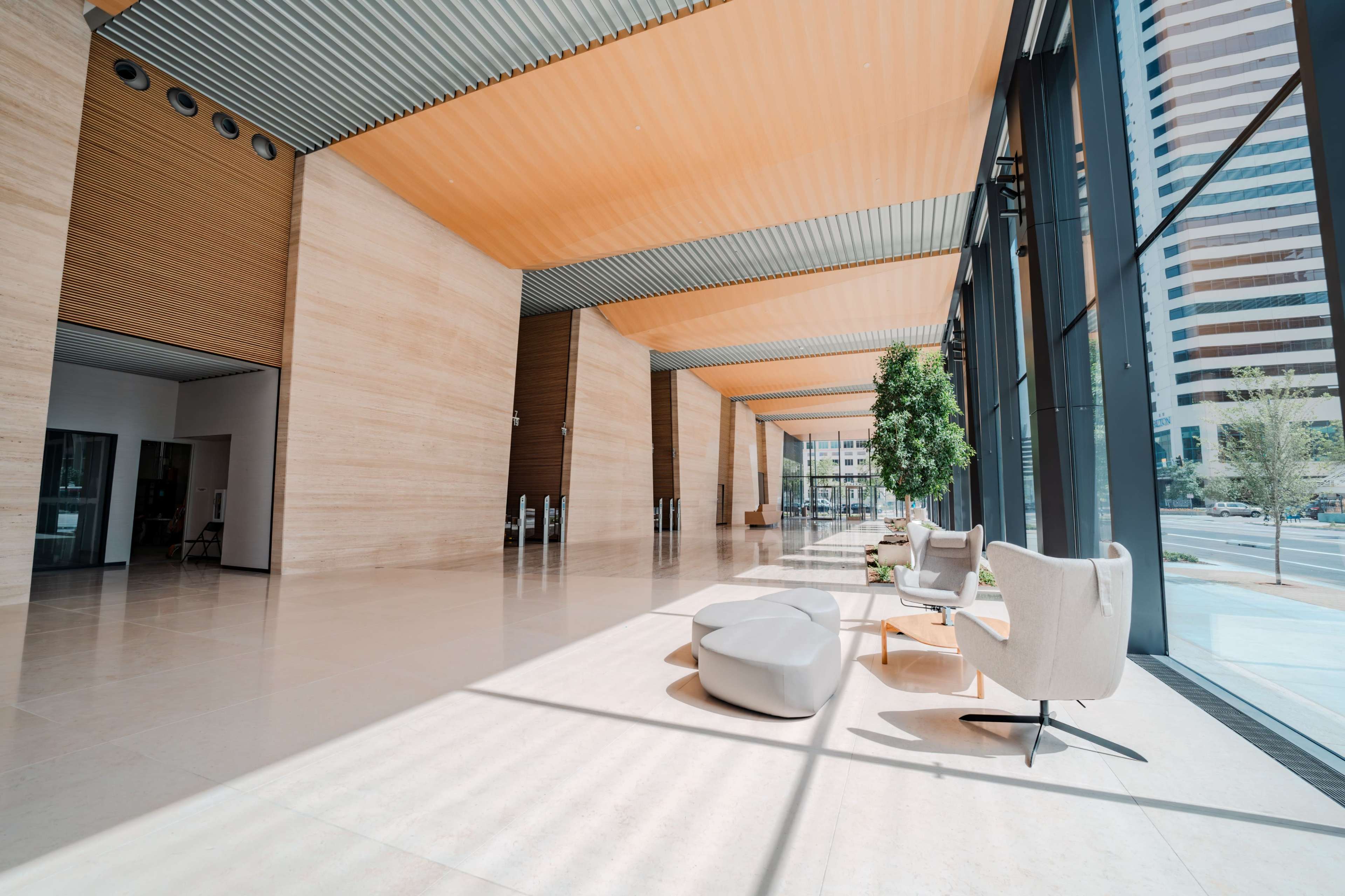 The image shows a spacious and modern lobby with large glass windows, minimalist furniture, and a leafy indoor plant.