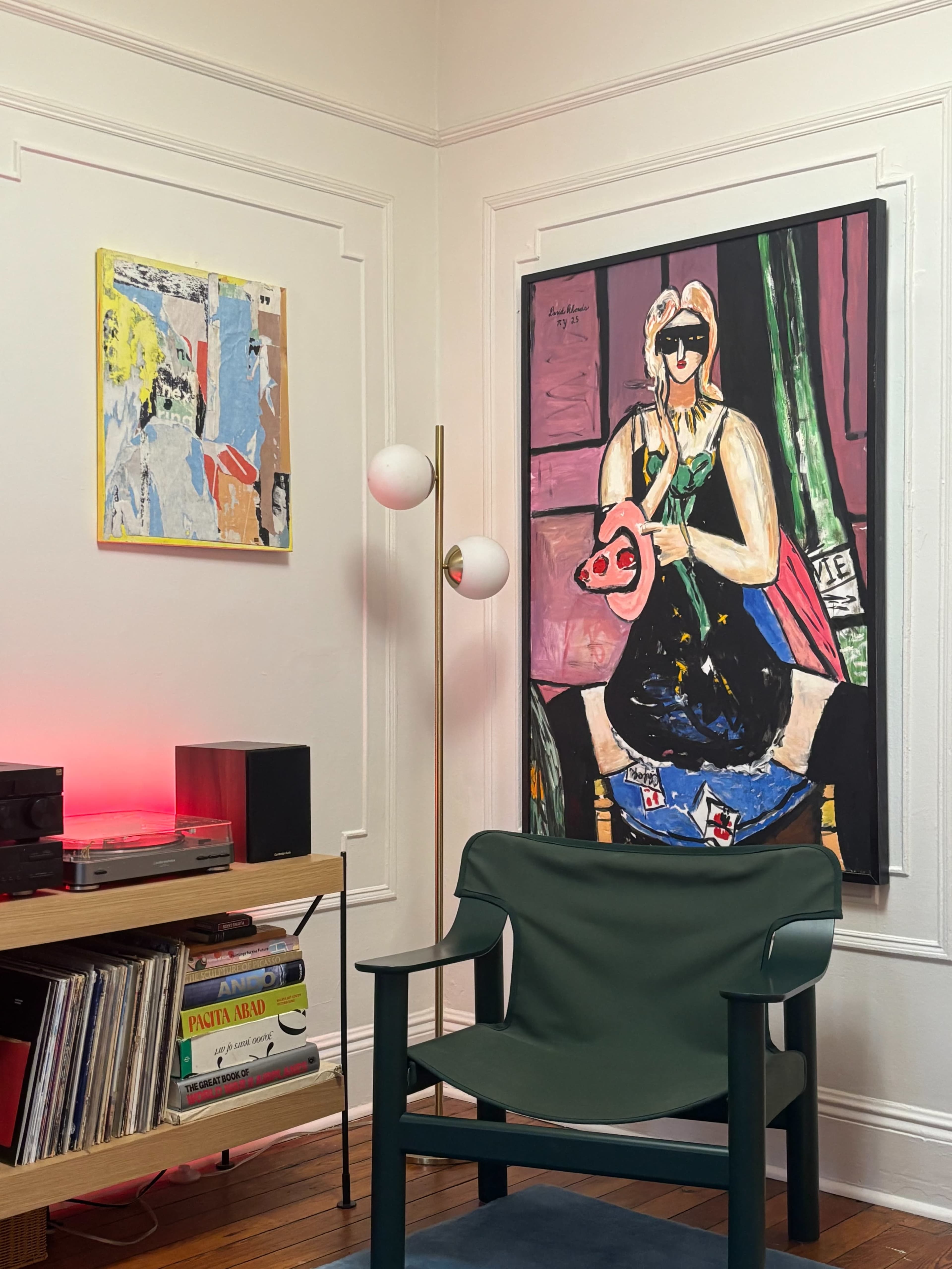 The scene features a corner of a room with colorful artwork hanging on the walls, a modern floor lamp, a green chair, and a record shelf filled with vinyl records.