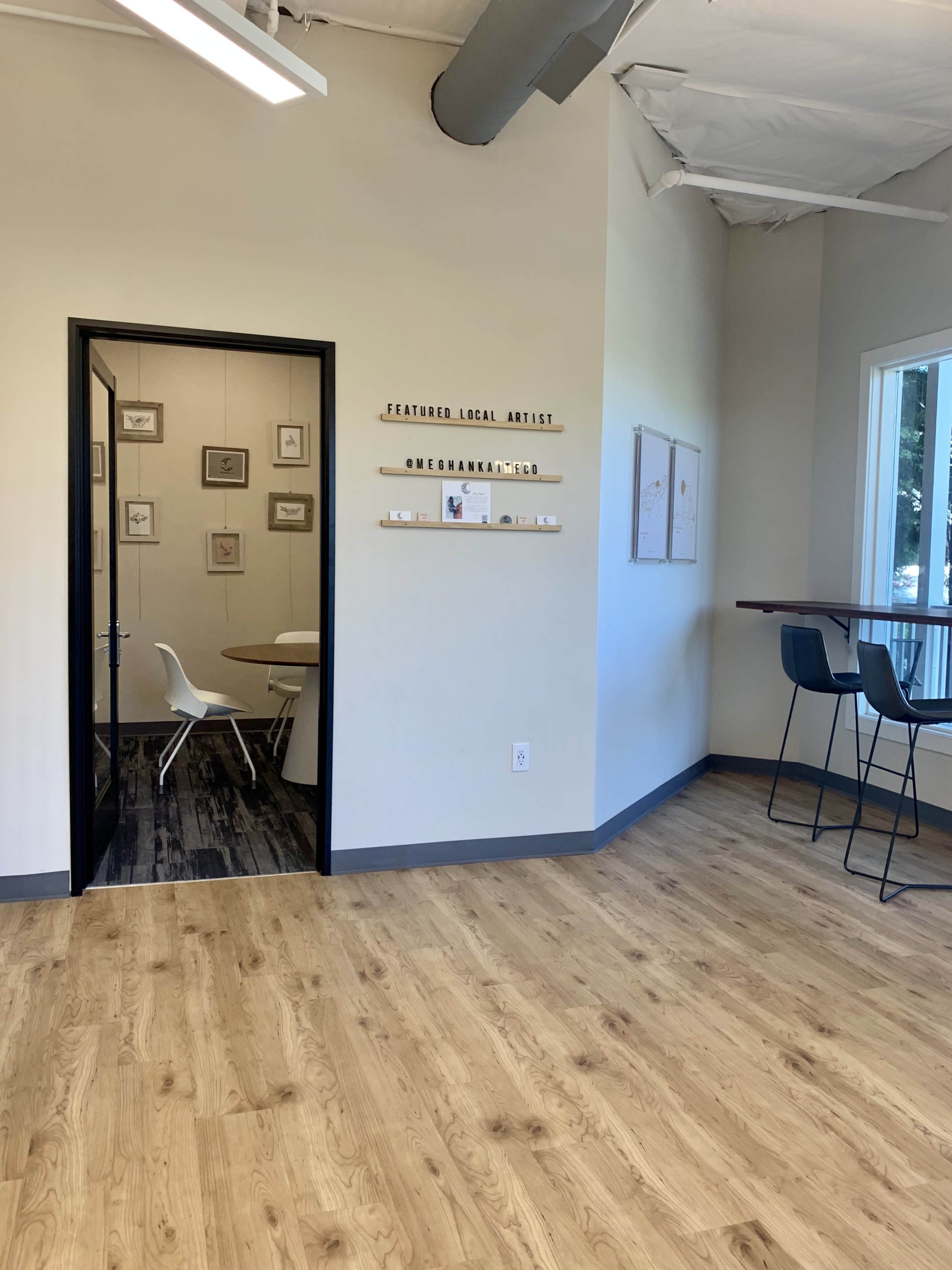 The image shows a minimalist room with a door leading to a meeting area, featuring a wooden floor and artwork displayed on the wall.
