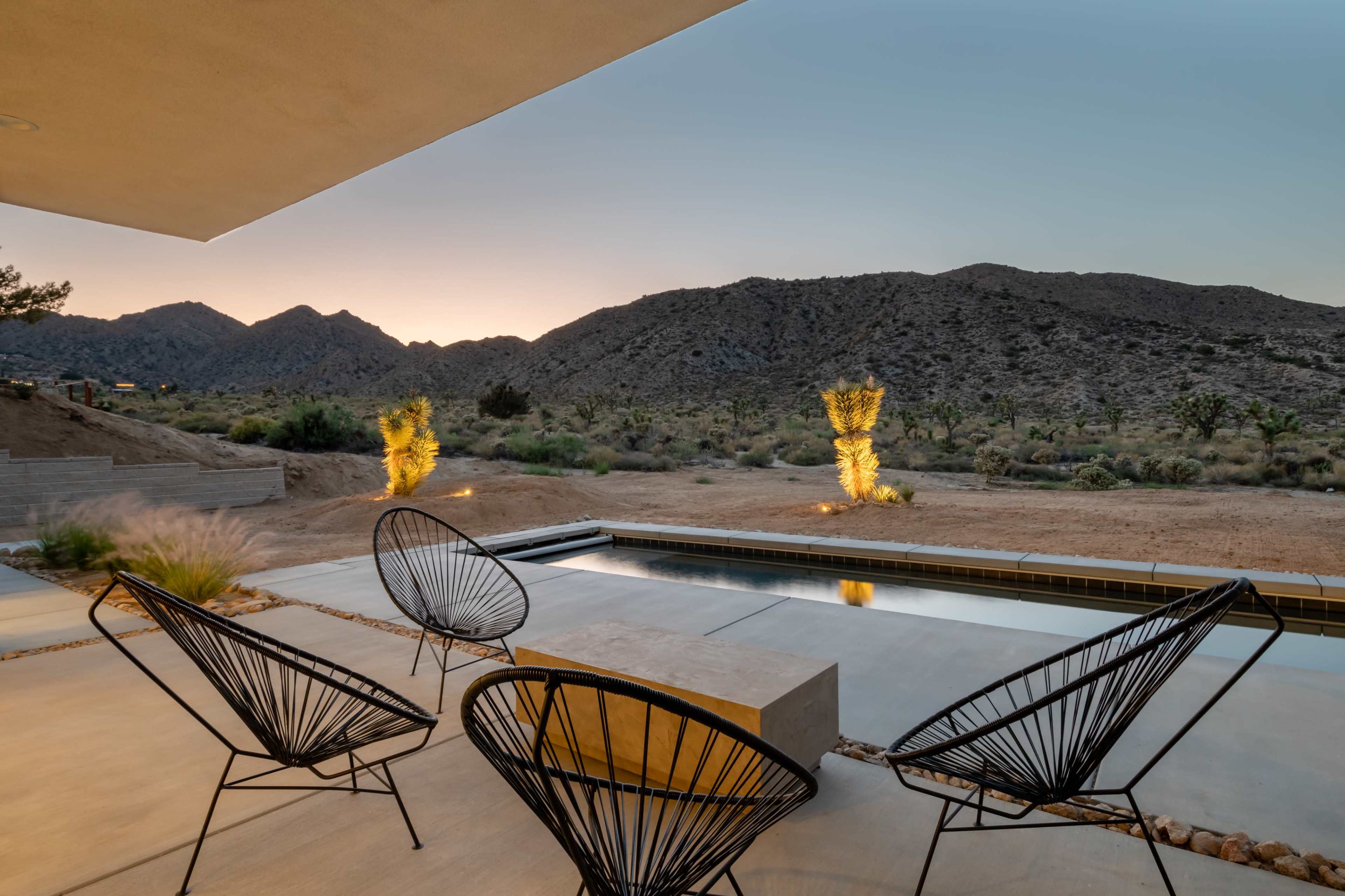 The image shows a modern patio with black chairs overlooking a desert landscape at sunset, framed by mountains in the background.