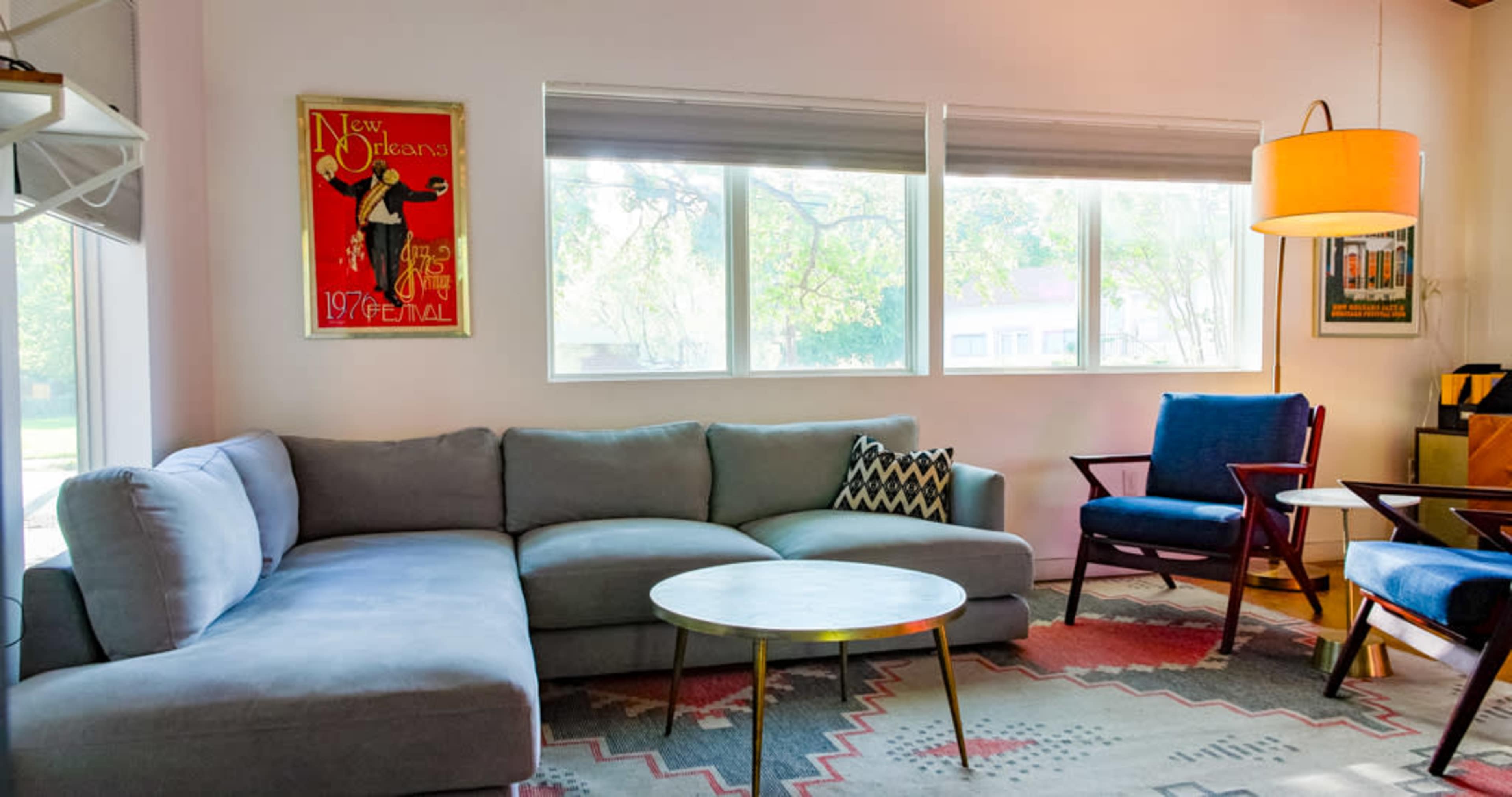 The image shows a modern living room featuring a gray sectional sofa, a round coffee table, and two accent chairs, with large windows letting in natural light and artwork on the walls.