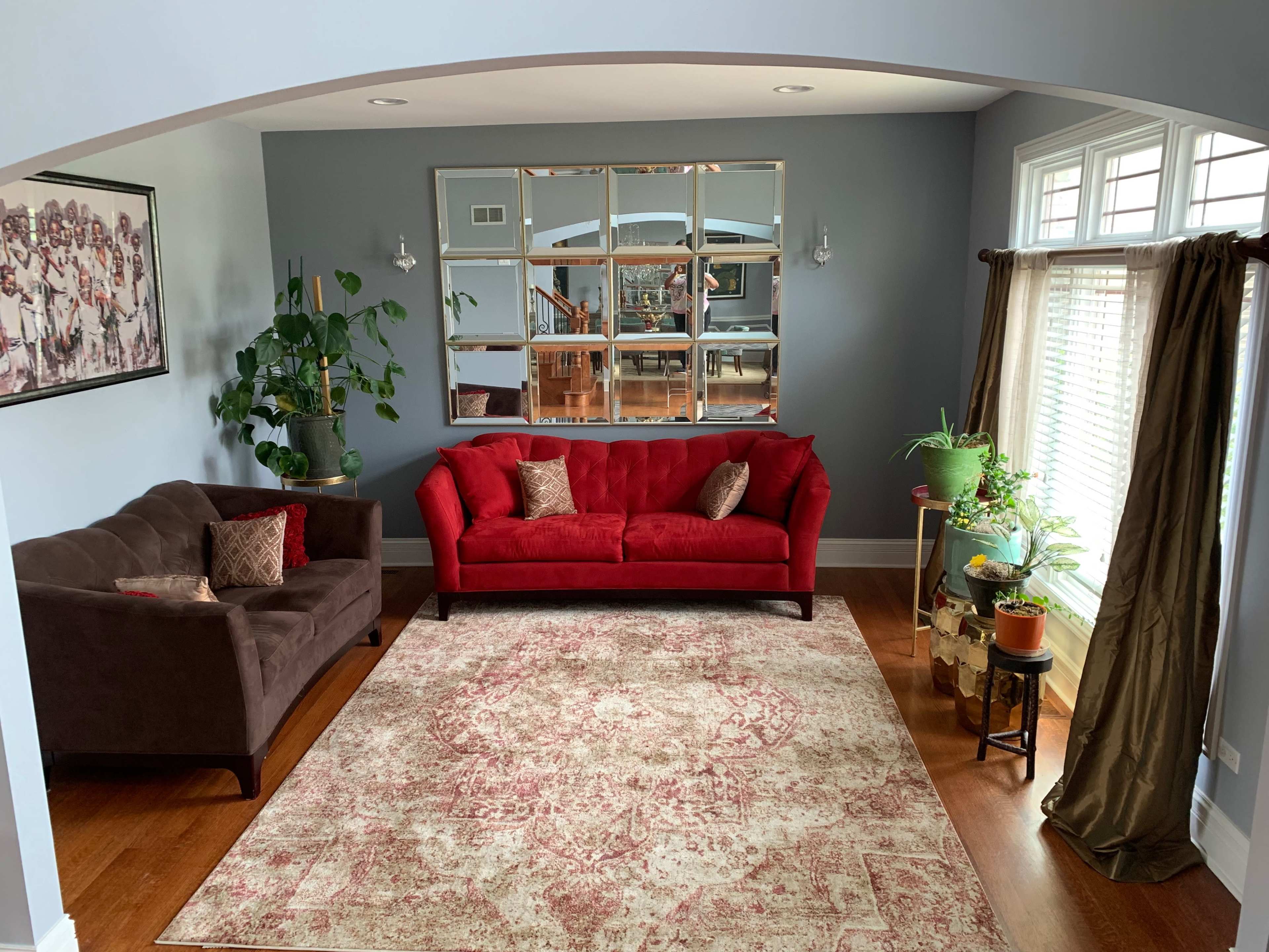 The living room features a red couch and a brown loveseat centered on a patterned area rug, surrounded by plants and framed mirrors against a blue wall.
