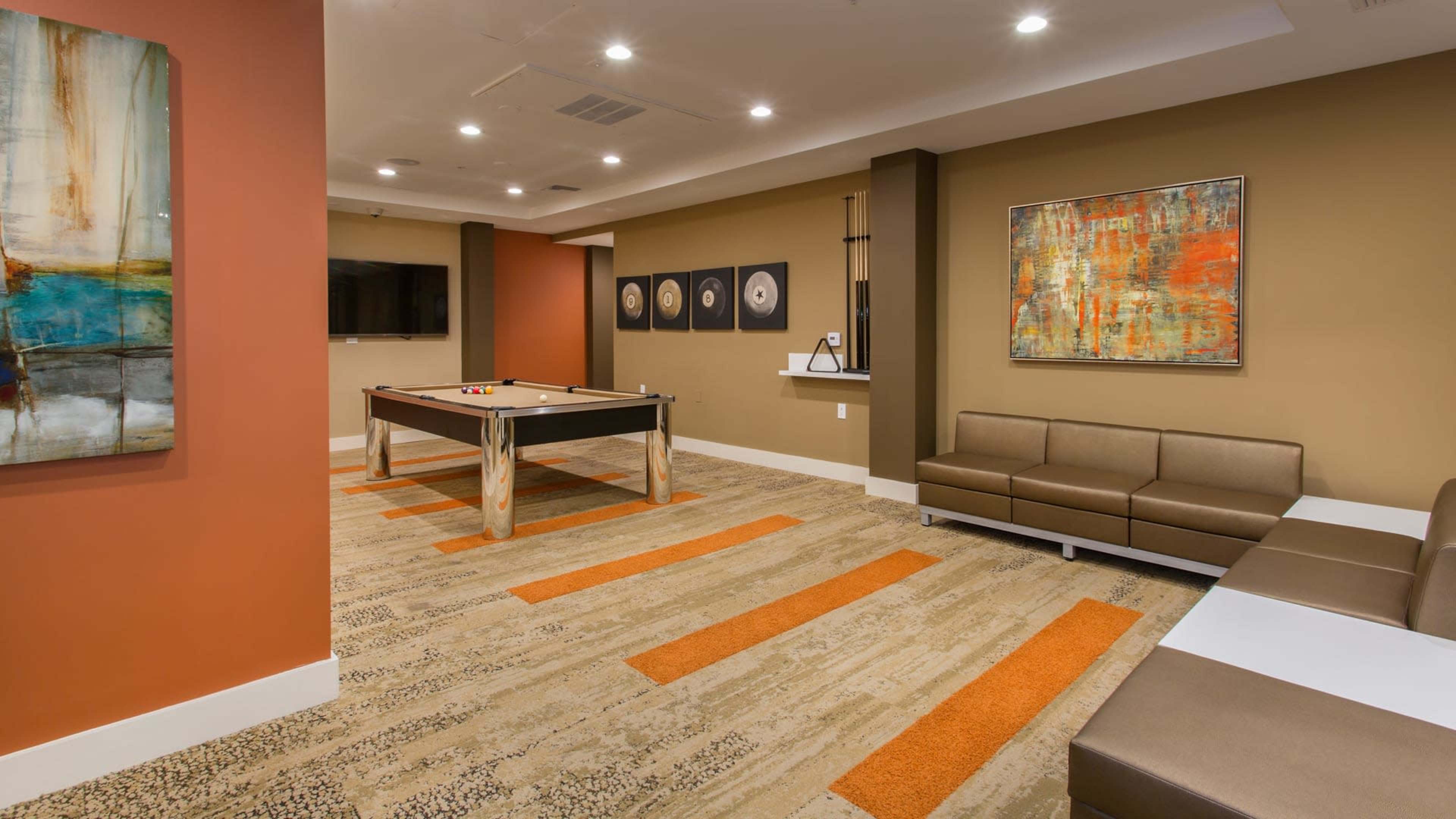 The image shows a modern lounge area featuring a billiard table, a sectional sofa, and abstract artwork on the walls.