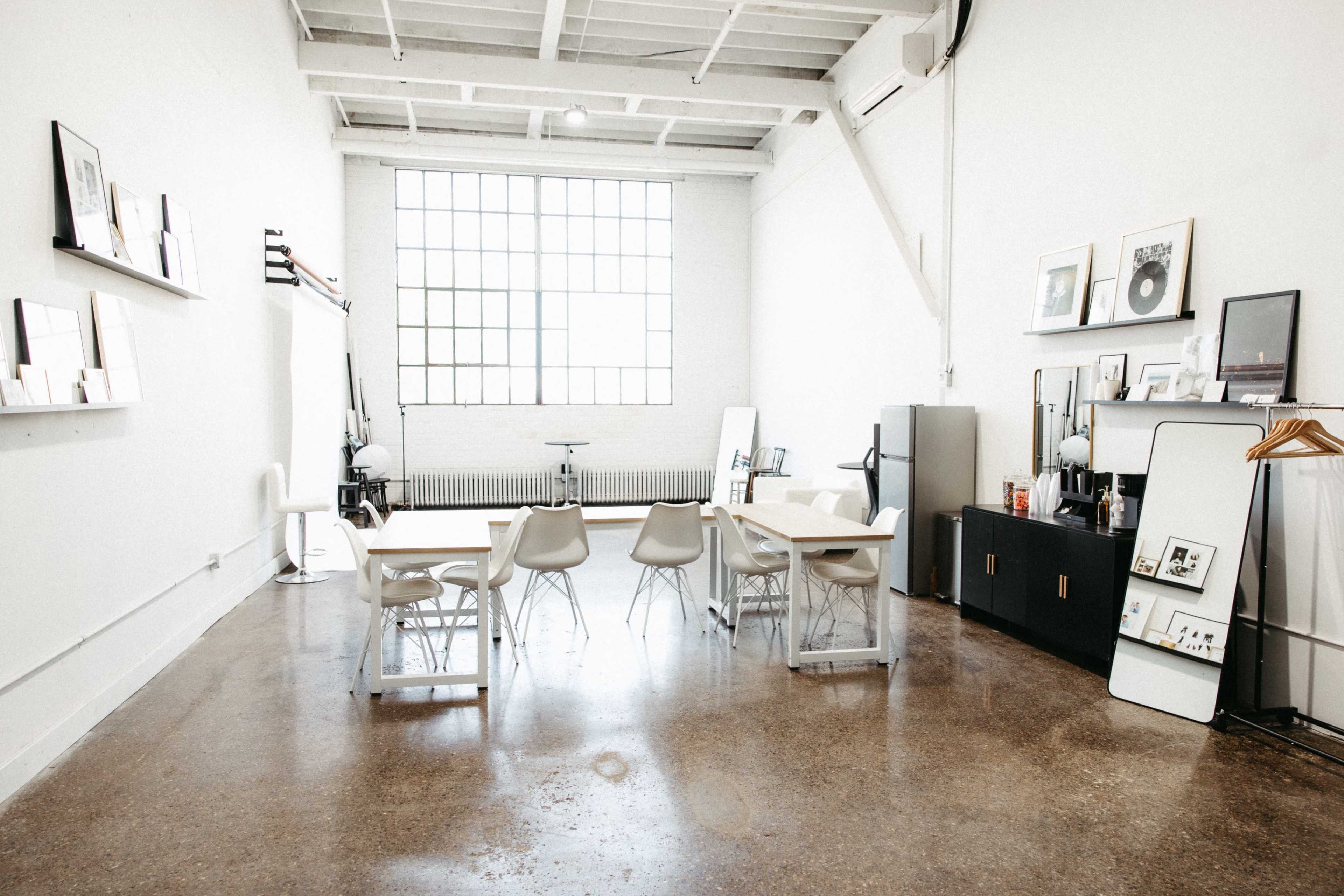 The image shows a spacious, minimalist studio with a large window, a long wooden table surrounded by four chairs, and various framed artworks on the walls.