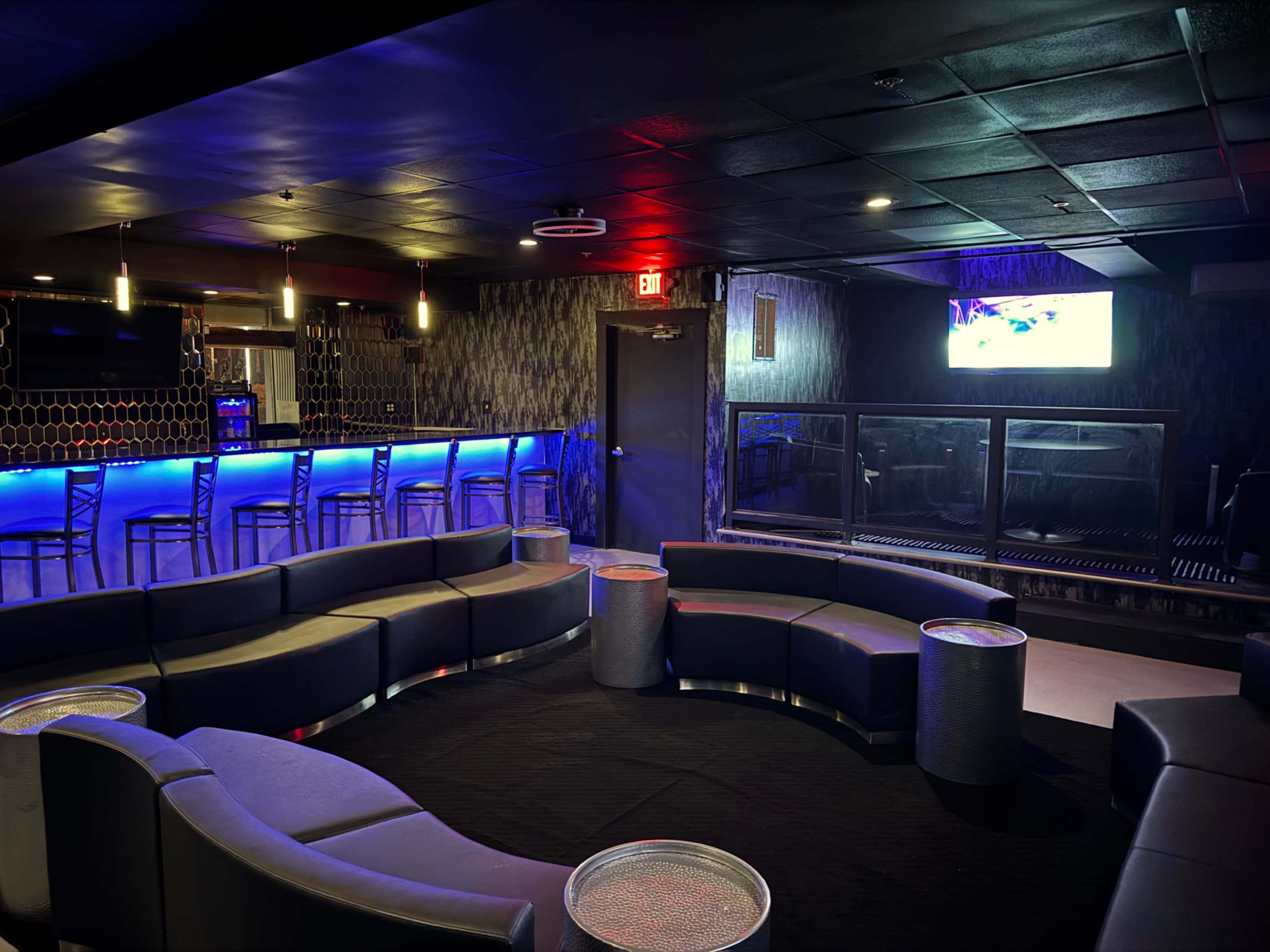 The image shows a modern lounge area featuring curved sofas, side tables, and illuminated blue bar with a television mounted on the wall.