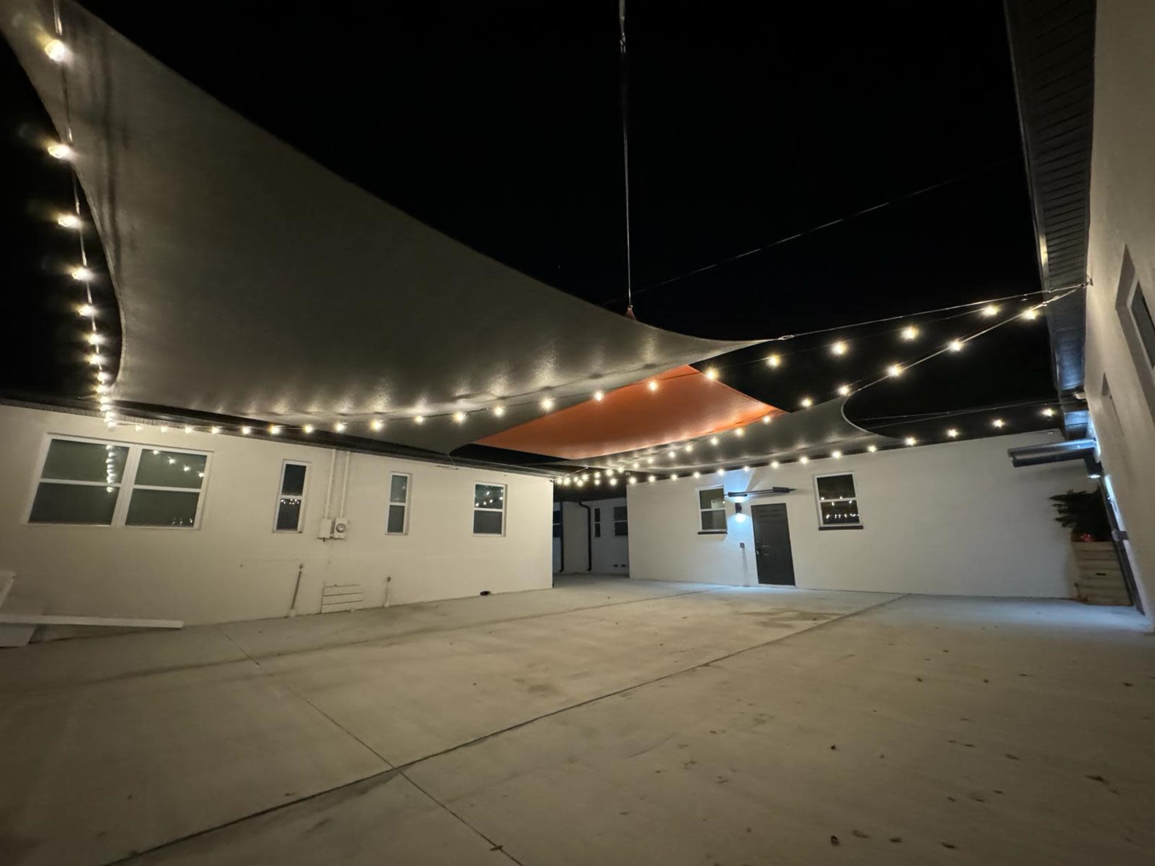 A well-lit courtyard at night with overhead fabric canopies and string lights suspended above a concrete surface, surrounding white walls and multiple doors.