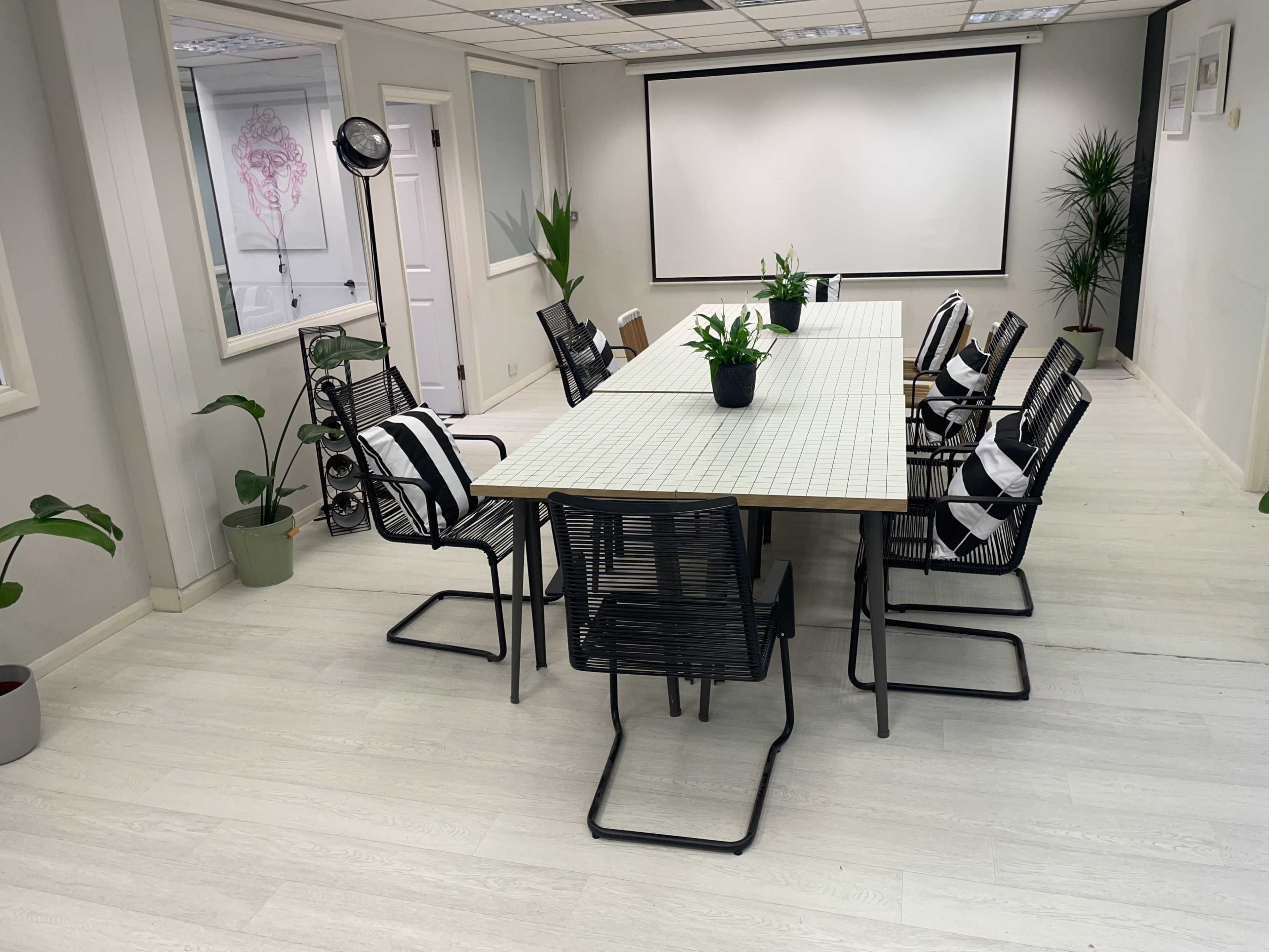 A modern conference room features a long, rectangular table surrounded by several black chairs, with potted plants in the corners and a projector screen at one end.