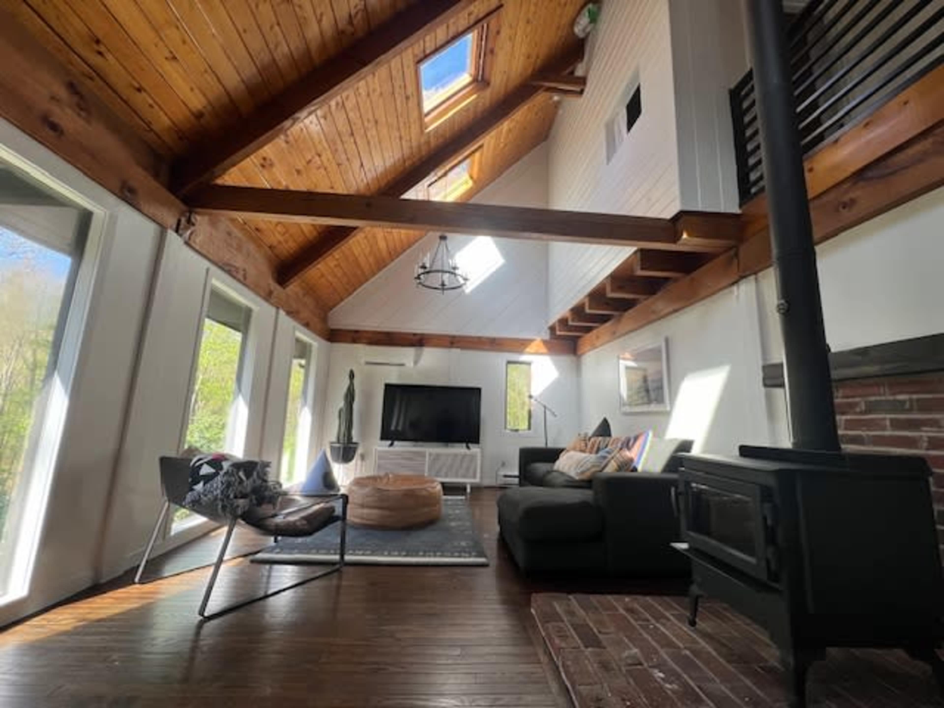 The living room features a high wooden A-frame ceiling with large windows, a television on the wall, a black sofa, and a wood stove.