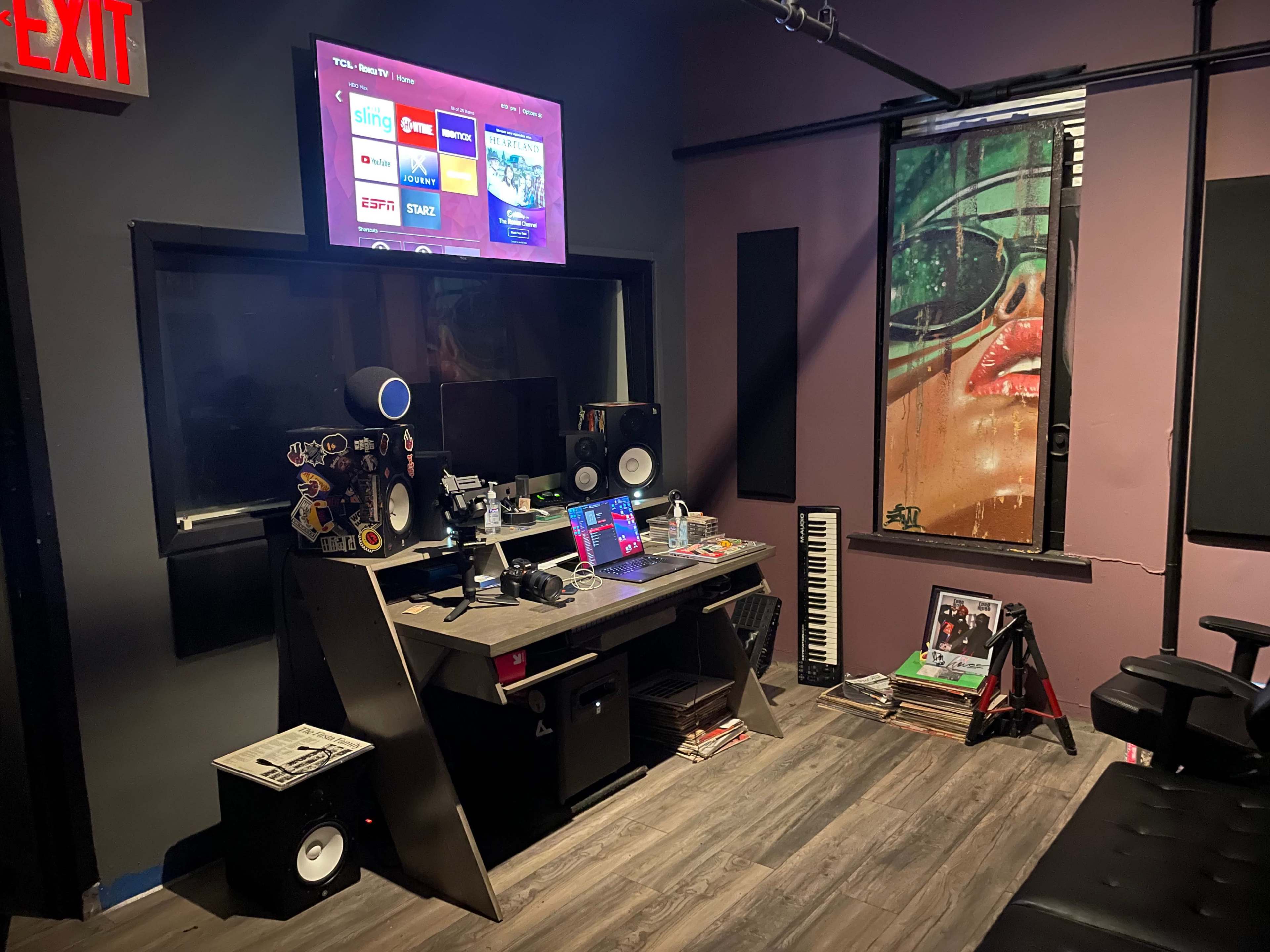 A music studio features a desk with audio equipment and a computer screen displaying streaming apps, alongside a painted mural and a keyboard on the floor.