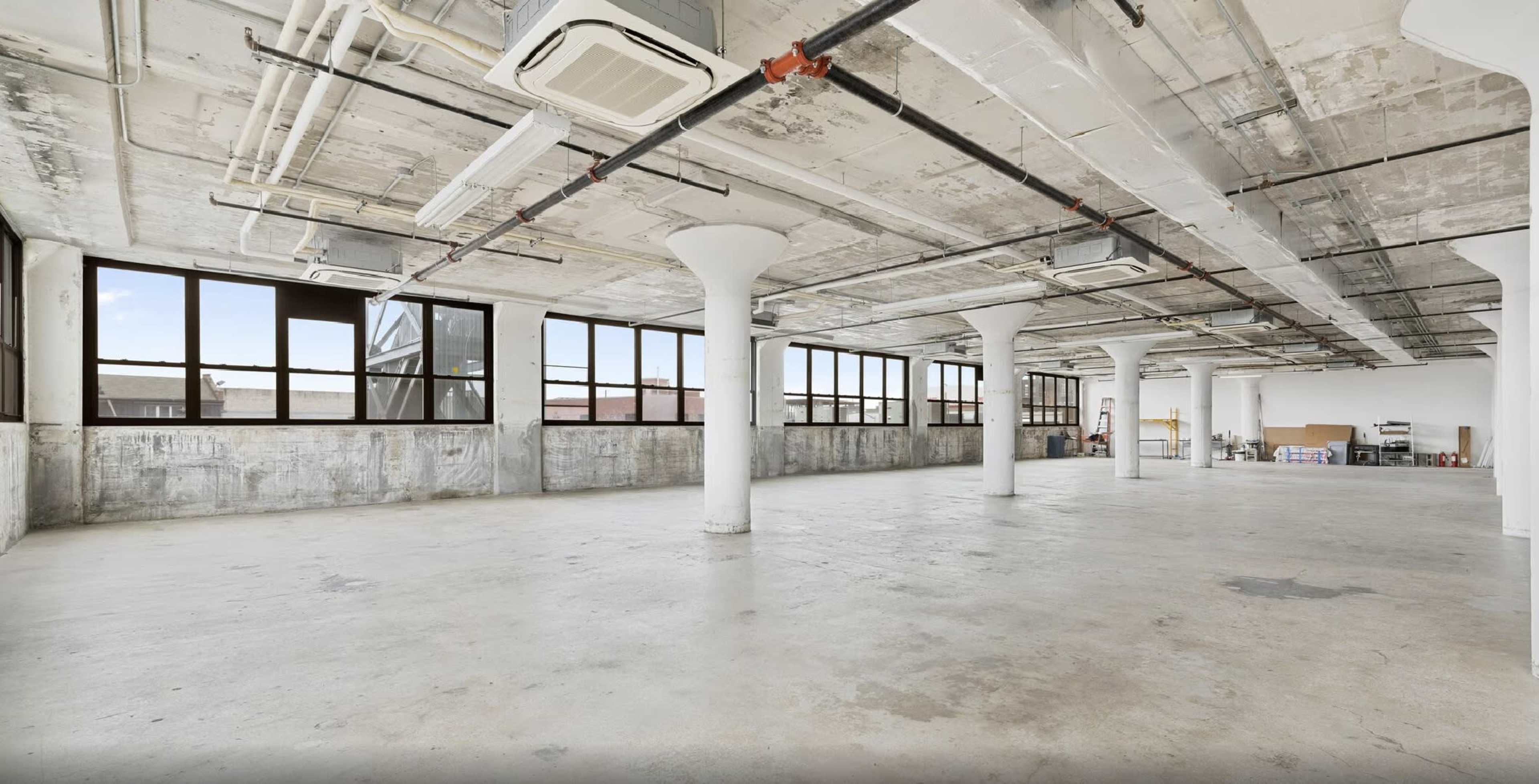 The image shows a spacious, industrial-style room with concrete floors, large windows, and exposed ceiling beams and pipes.