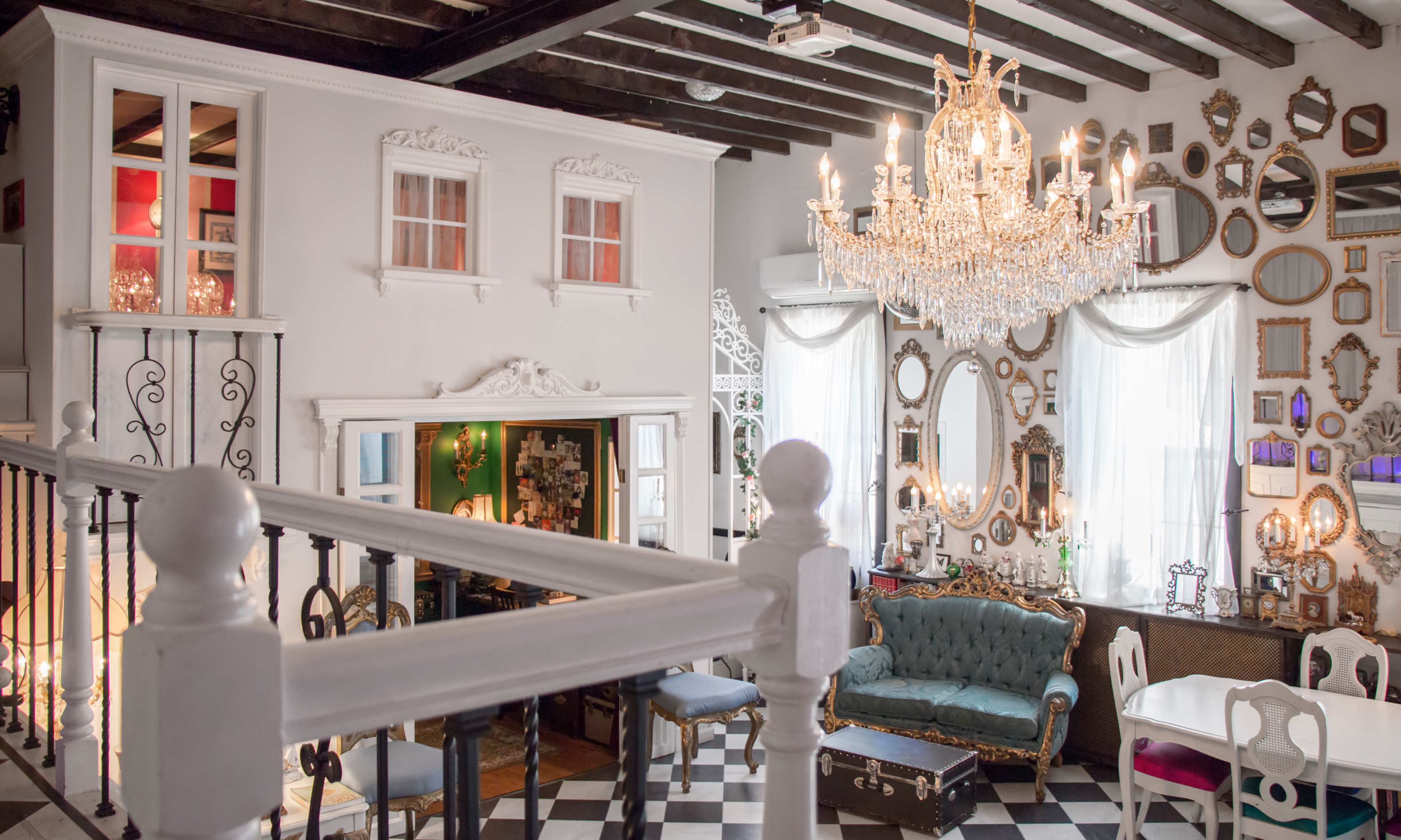 An elegantly designed interior features a vintage sofa, a crystal chandelier, and numerous framed mirrors on the walls, with a staircase leading to a small balcony.
