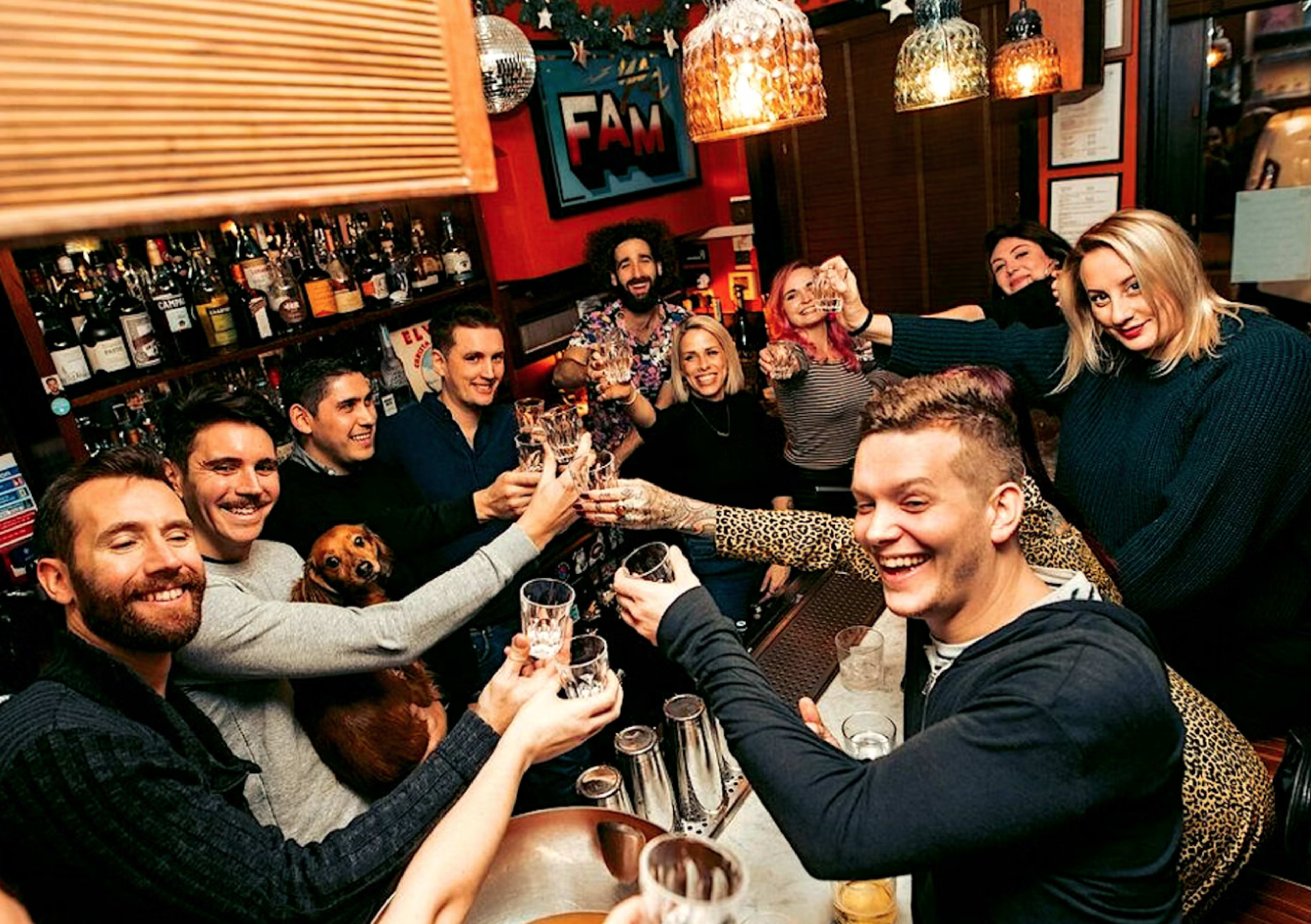 A group of people is gathered around a bar, raising glasses in a toast while a dog sits among them.